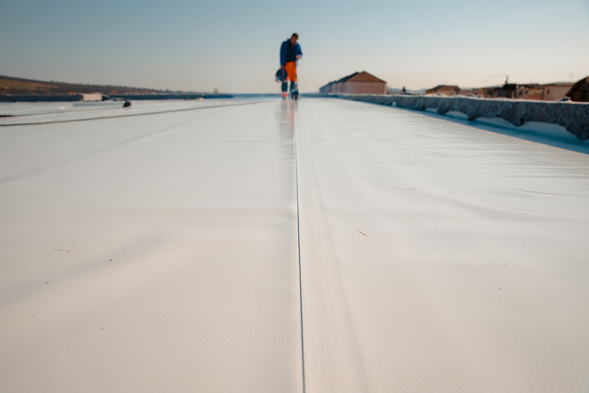 Worker installs white waterproof membrane on a flat rooftop under a clear sky.