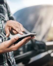 Person with a phone, standing by a car with an open hood, outdoors.