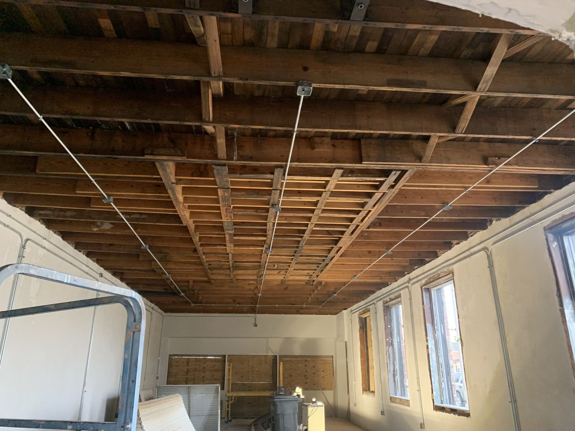 Interior view of a room with exposed wooden ceiling beams and support wires. Walls are light-colored.