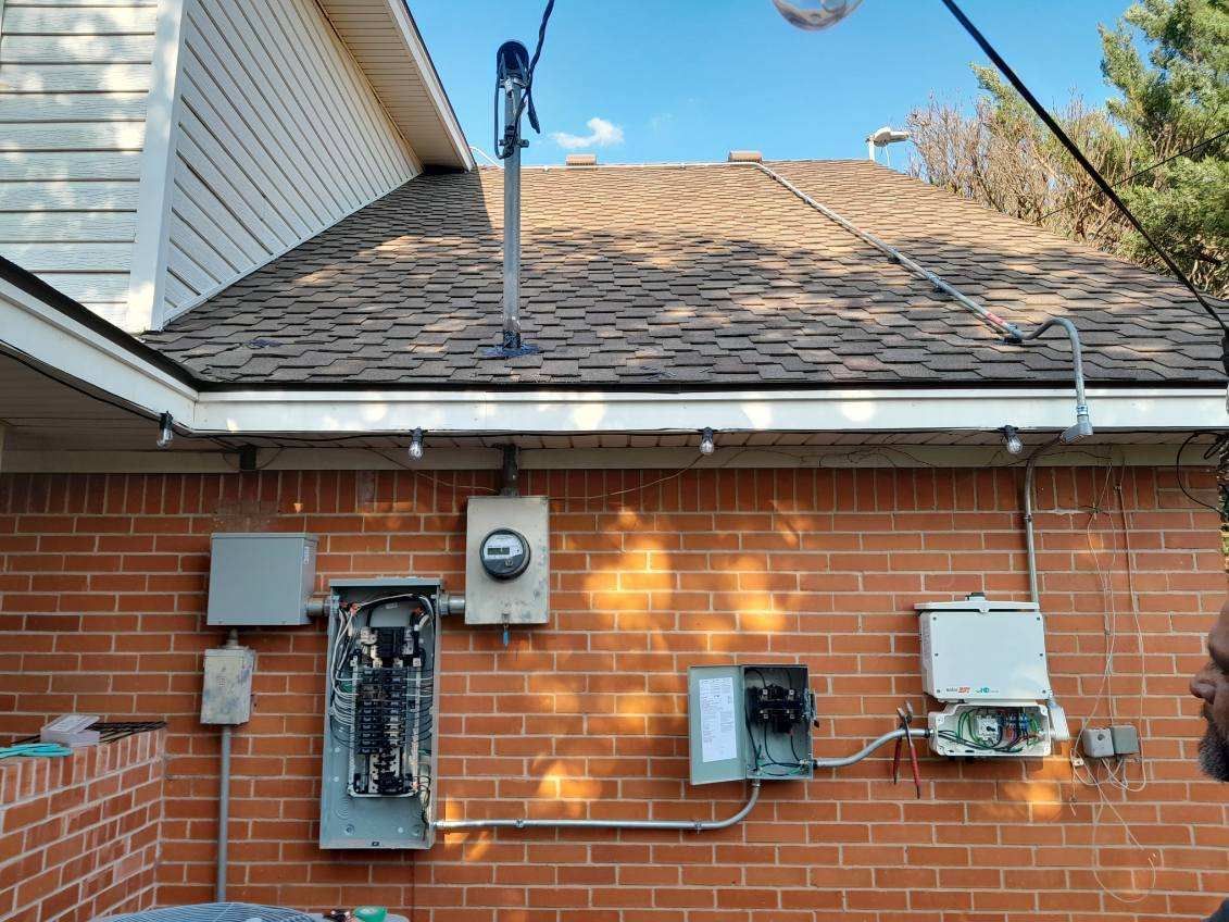 Electrical equipment on a brick wall: panel, meter, and boxes beneath a roof with overhead wires.