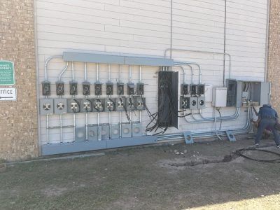 Electrical meters and conduit on building exterior; worker near open panel. Gray and tan.