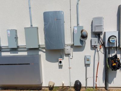 Outdoor electrical components mounted on a cream-colored wall: a large battery, several boxes, conduit, and a utility meter.