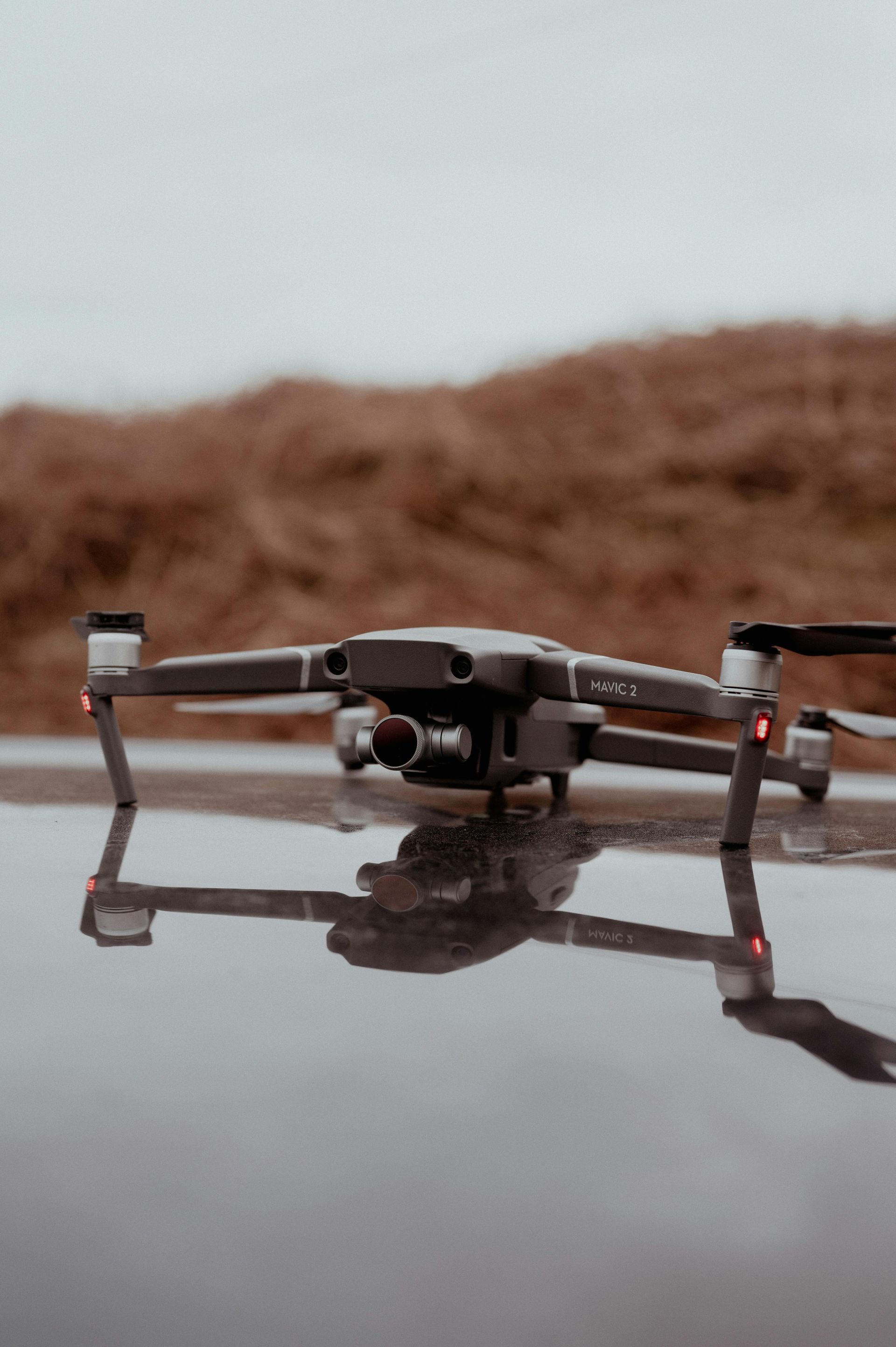 Drone with rotors extended, reflected in wet ground, against a blurred brown hill under a cloudy sky.