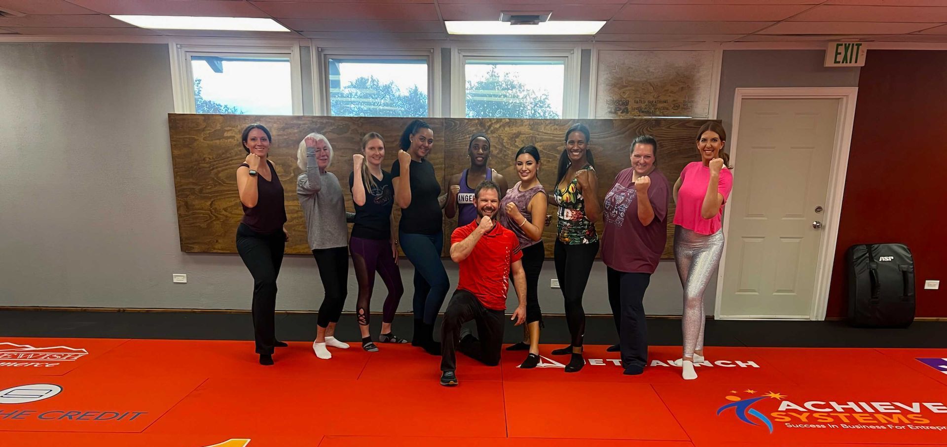 Group of people, likely a fitness class, posing with fists up on a red mat. They are indoors, with a decorative wall, windows, and an