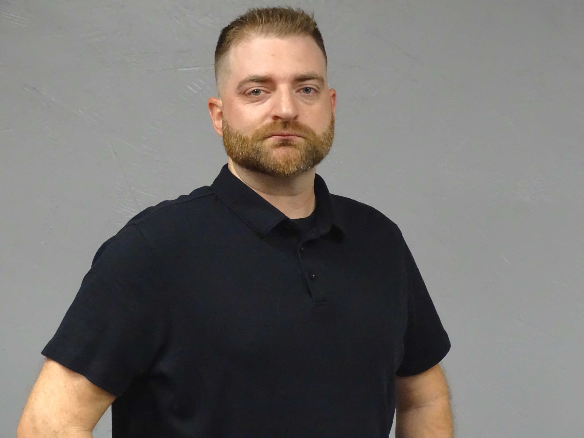 A man with a beard is wearing a black polo shirt.