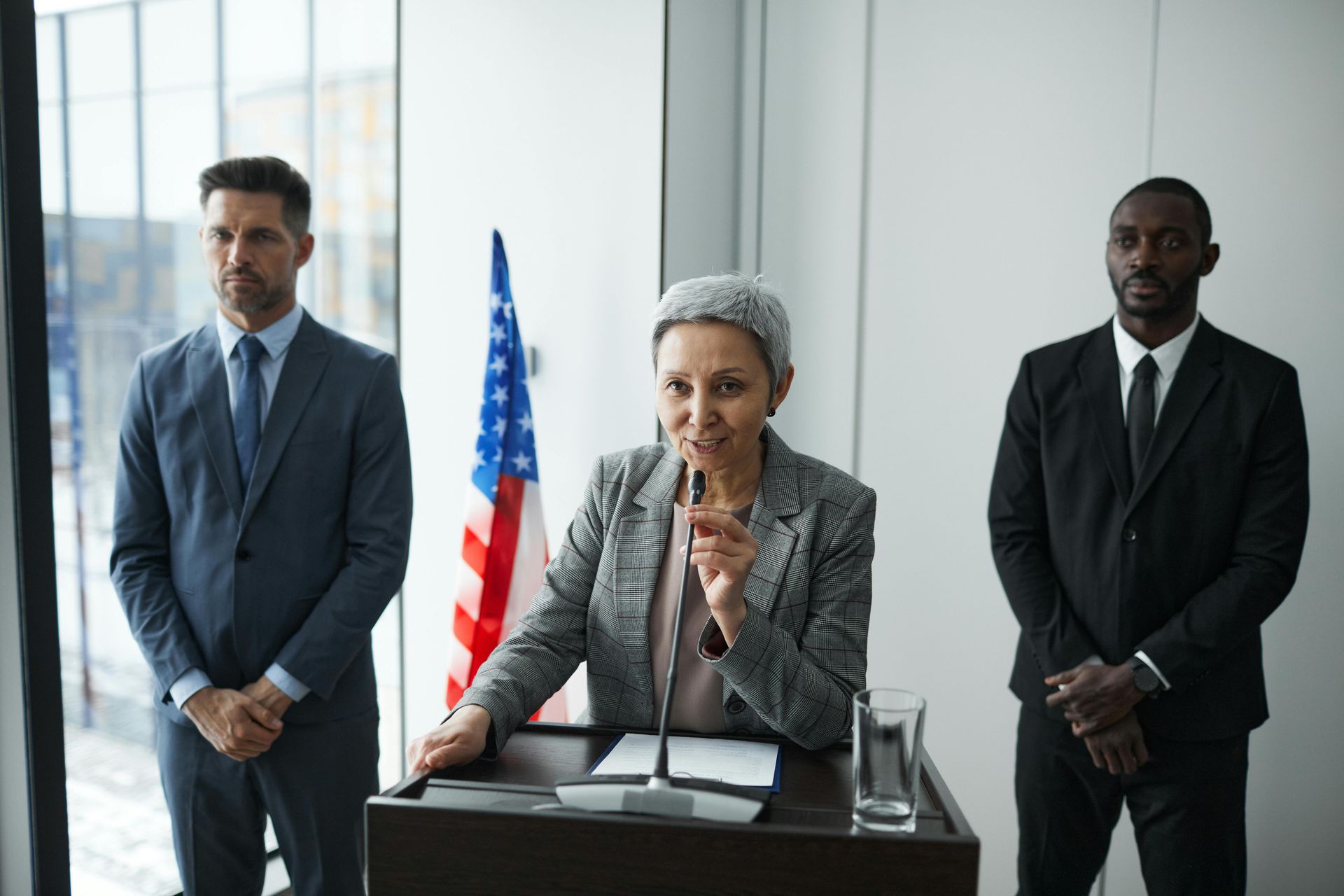 A woman is giving a speech at a podium with two men standing behind her.
