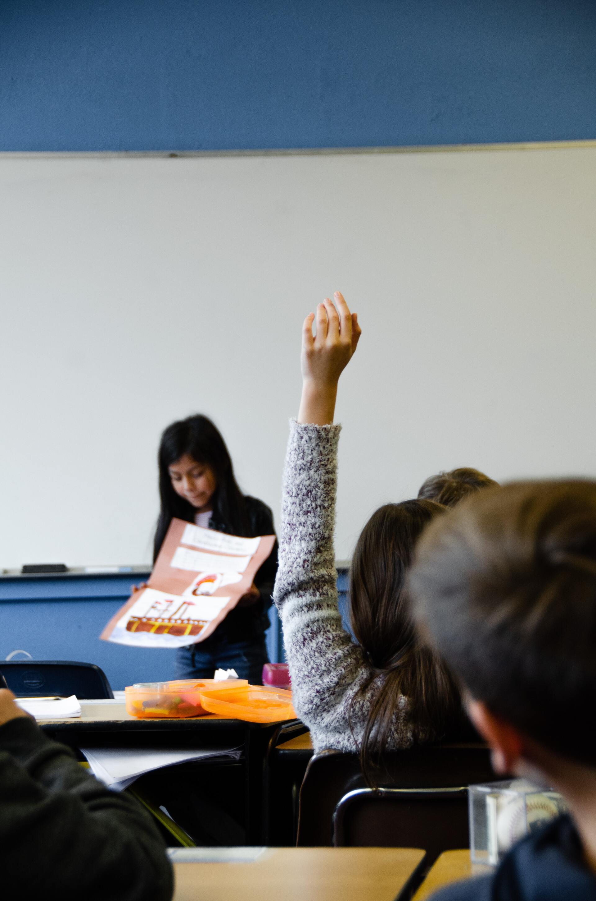 A girl is raising her hand in a classroom to answer a question.
