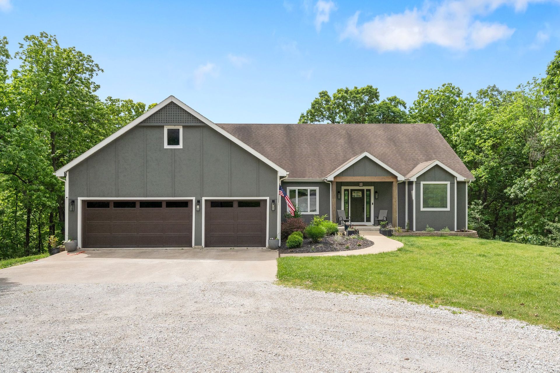 Gray house with brown garage doors and a gravel driveway, surrounded by green trees.