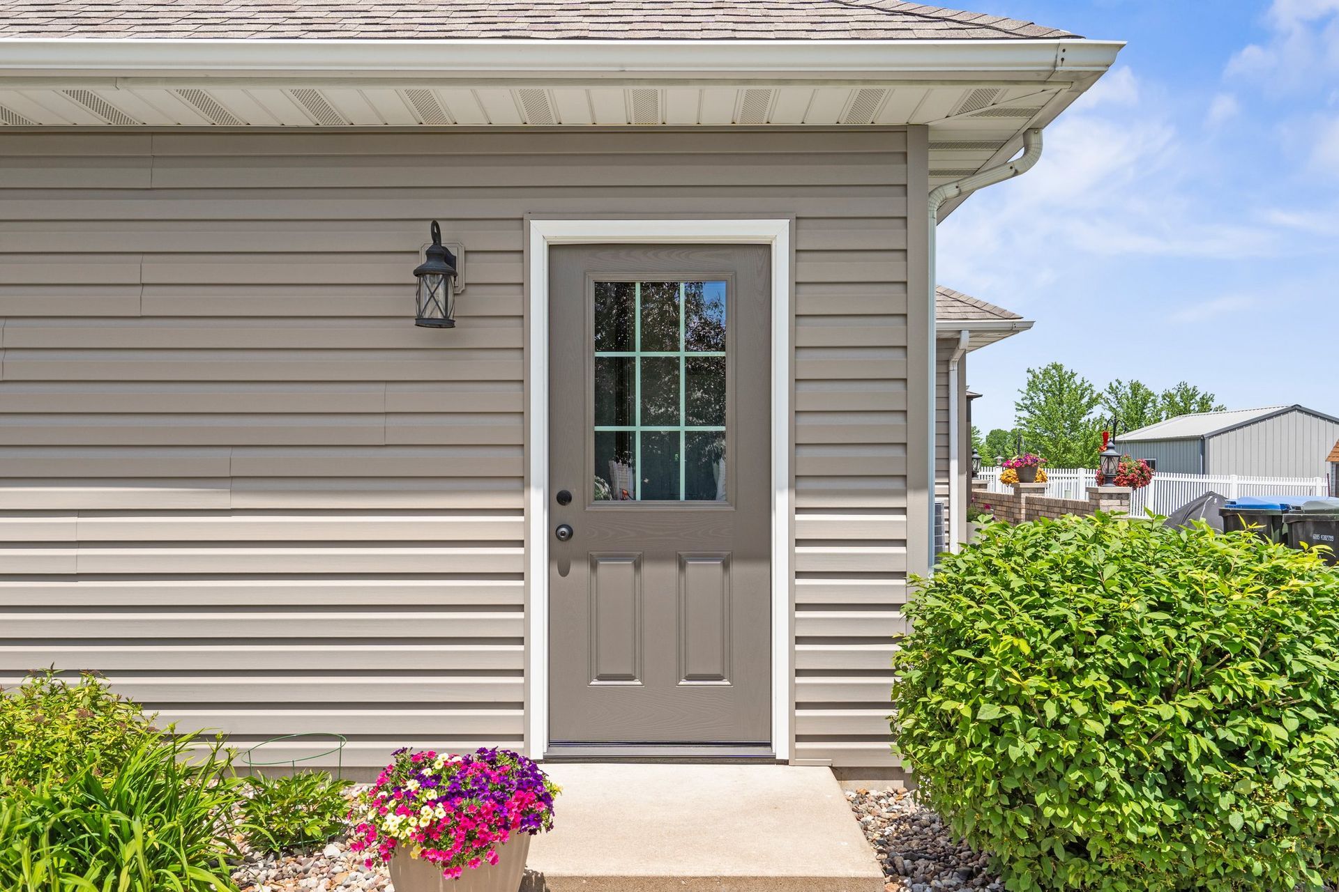 Tan door with window, surrounded by gray siding, lit by a black lantern, with a small bush and flower pot in front.