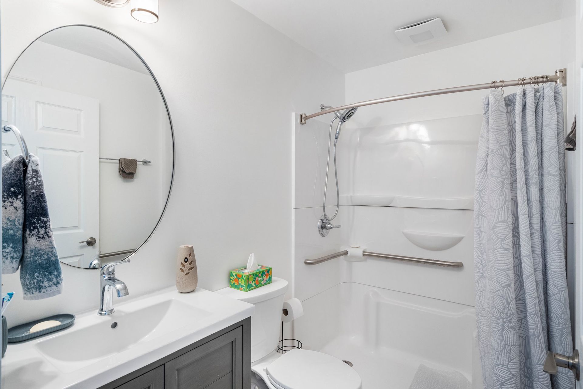 White bathroom with gray vanity, oval mirror, shower with curtain, and a towel rack.