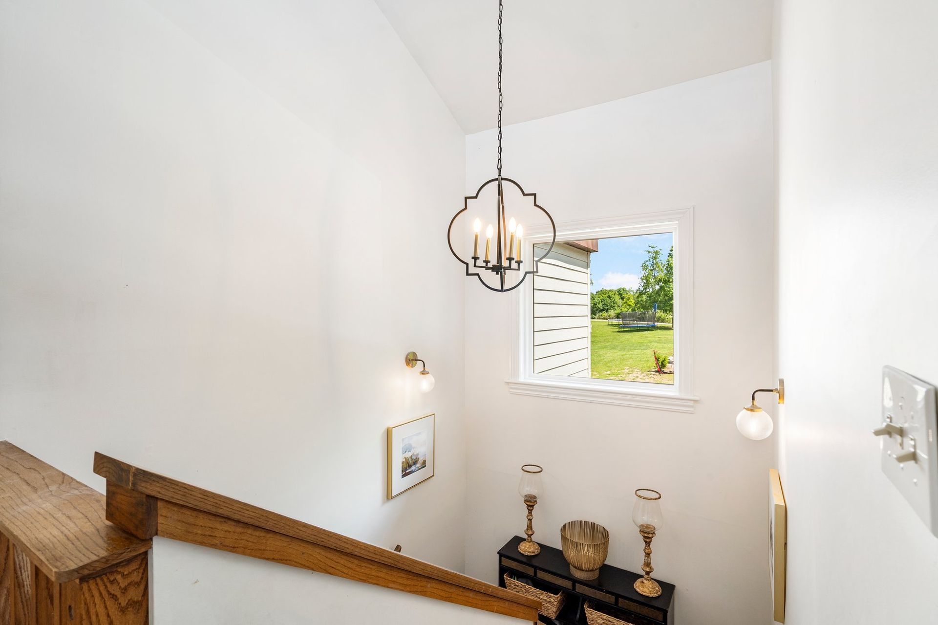 Interior view of a stairwell with a chandelier, window overlooking a yard, and wall sconces.