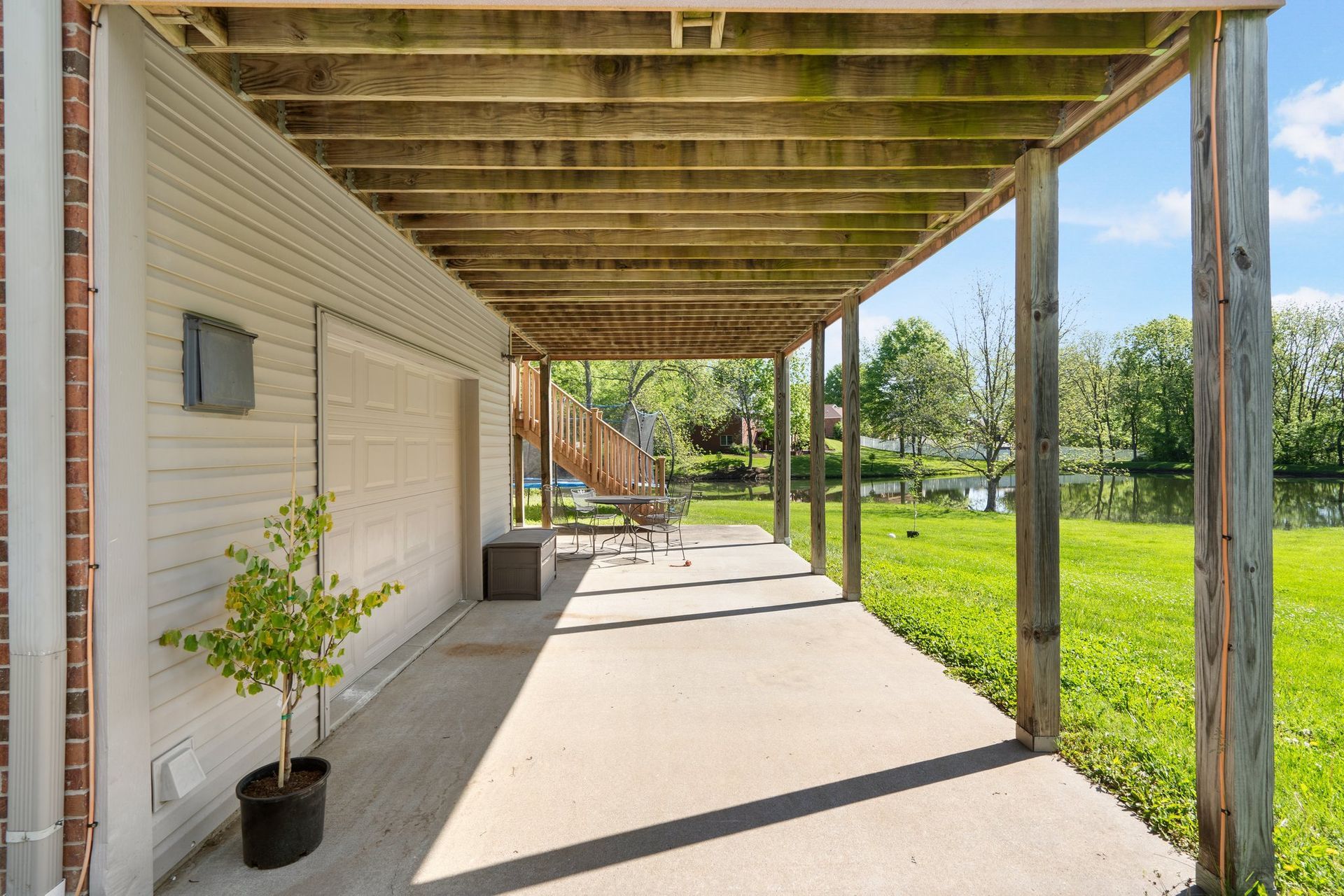 Covered concrete patio overlooking a grassy area and water; wooden beams and a potted tree are present.