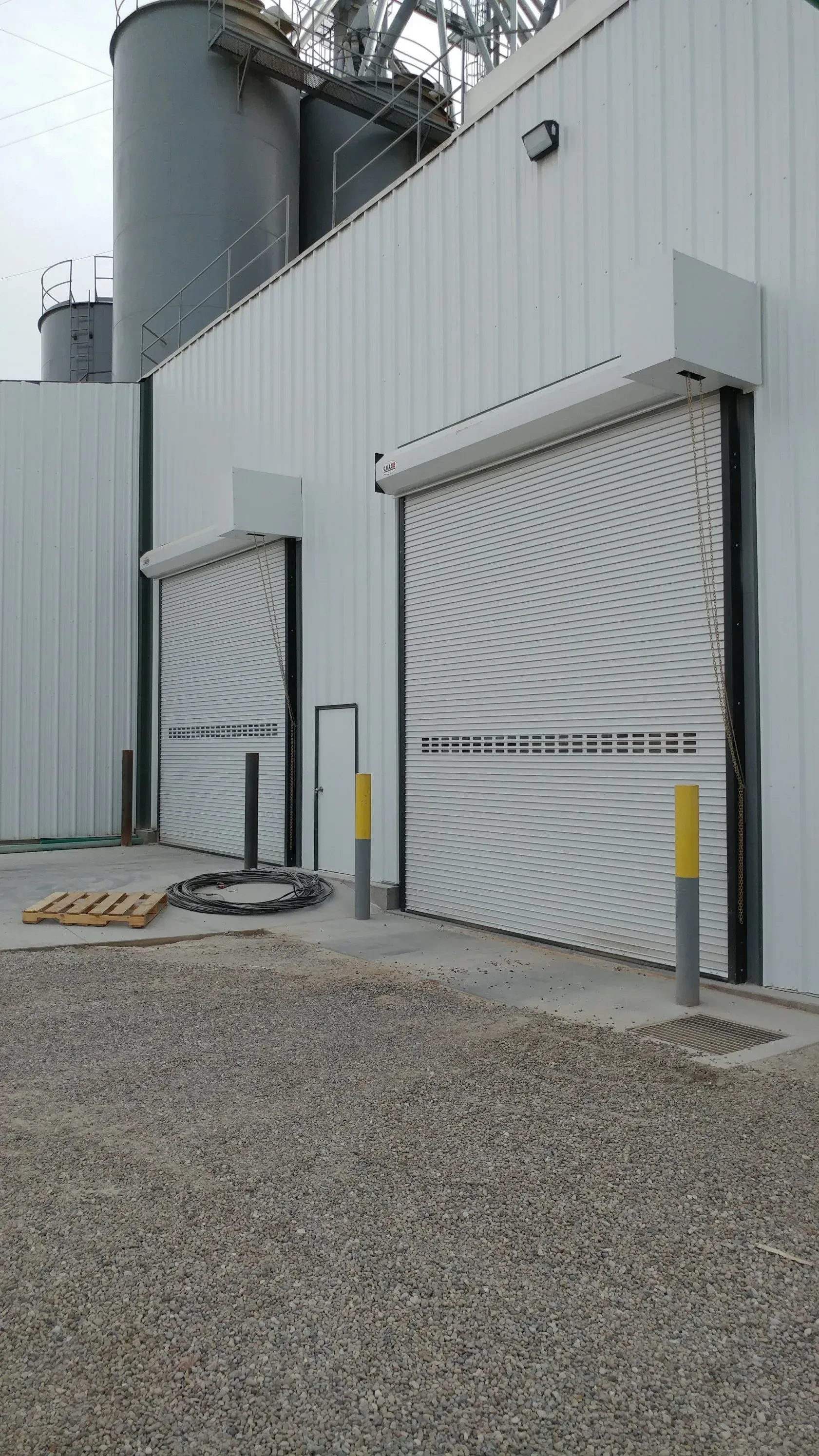 White industrial building with two large, closed roll-up doors; gray gravel ground.