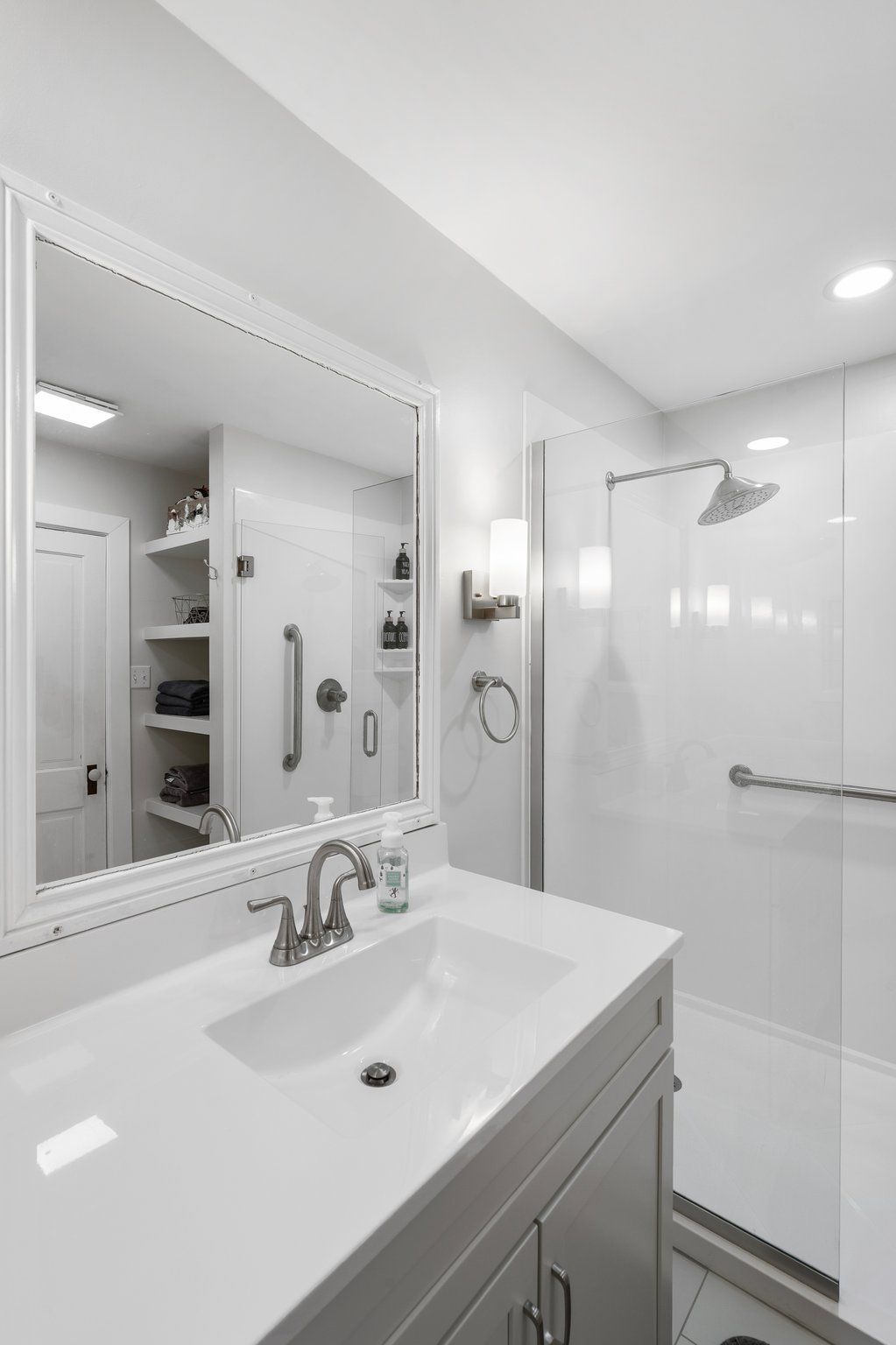 White bathroom with accessible features: vanity, shower, grab bars, open shelving, and a large mirror.