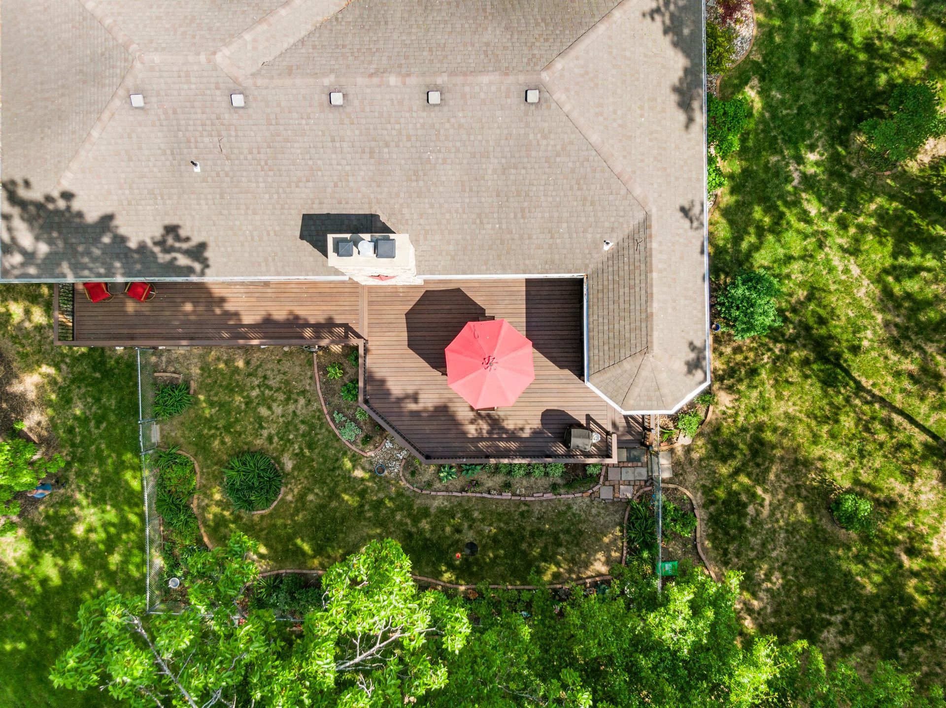 Overhead view of a house with a brown roof and a wooden deck with a red umbrella; surrounded by green grass and trees.