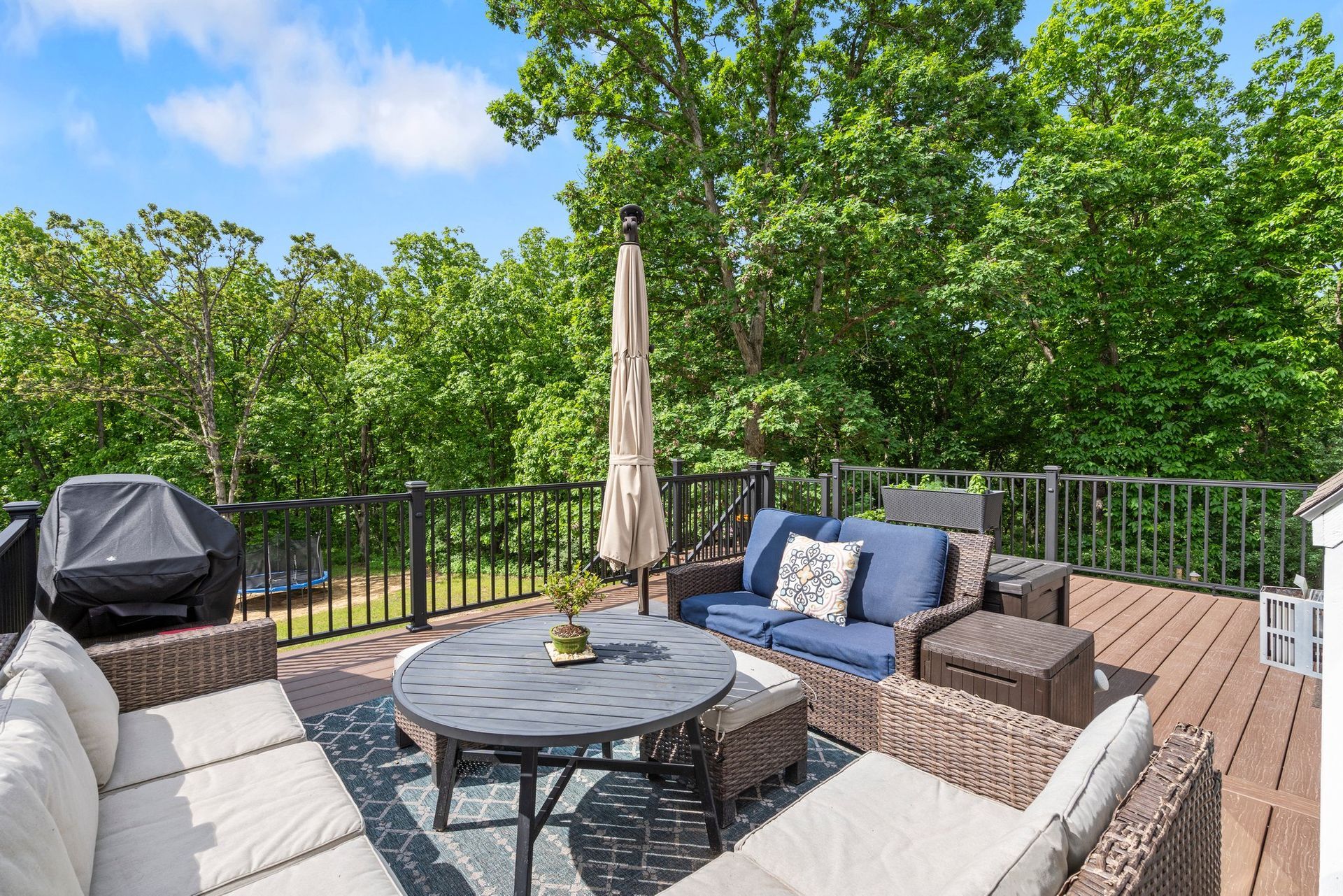 Deck with outdoor furniture: sofas, table, umbrella, grill. Lush green trees in the background, blue sky.