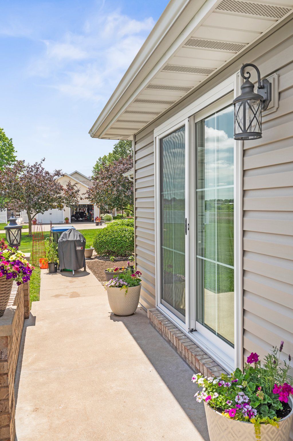 Sliding glass doors on a beige house, with a walkway lined with potted flowers, overlooking a green lawn.