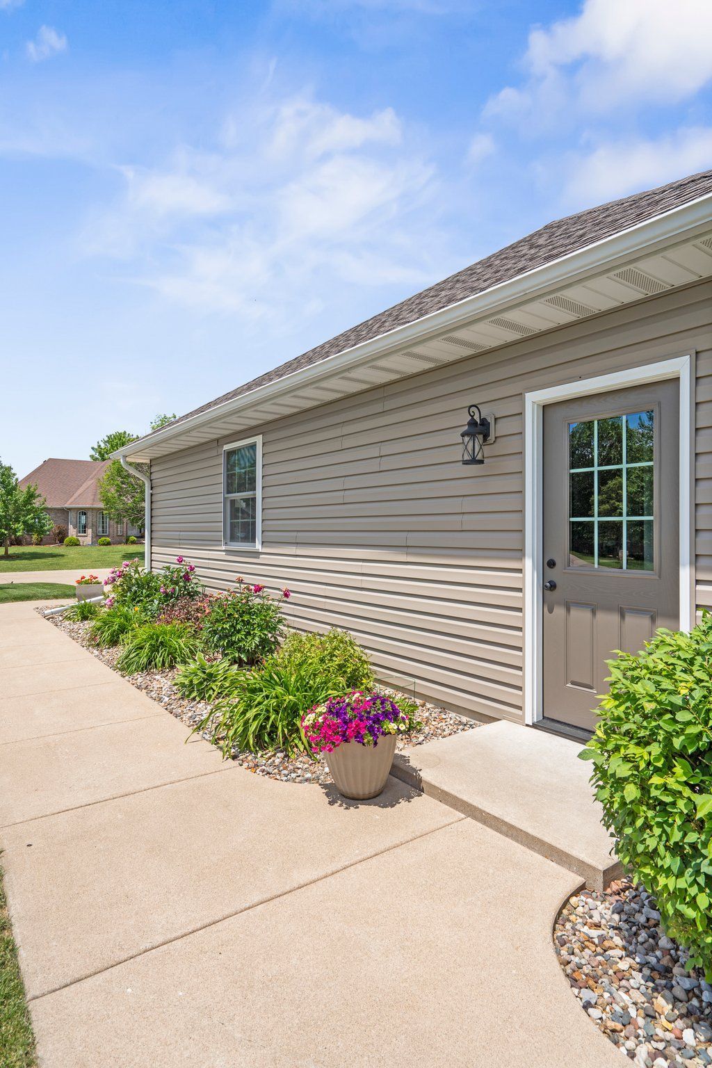 Tan house with a concrete walkway, garden bed, and a brown door under a bright sky.