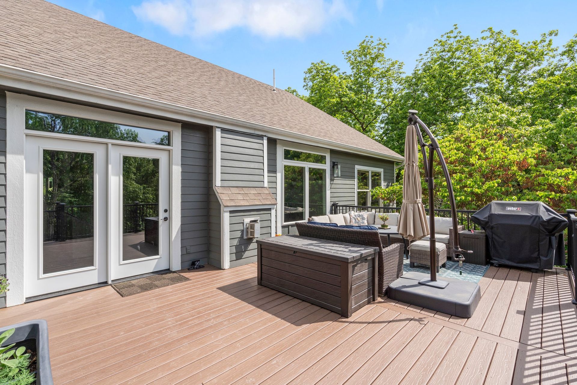 A deck with outdoor furniture next to a house with large windows. The sky is blue and trees surround the house.