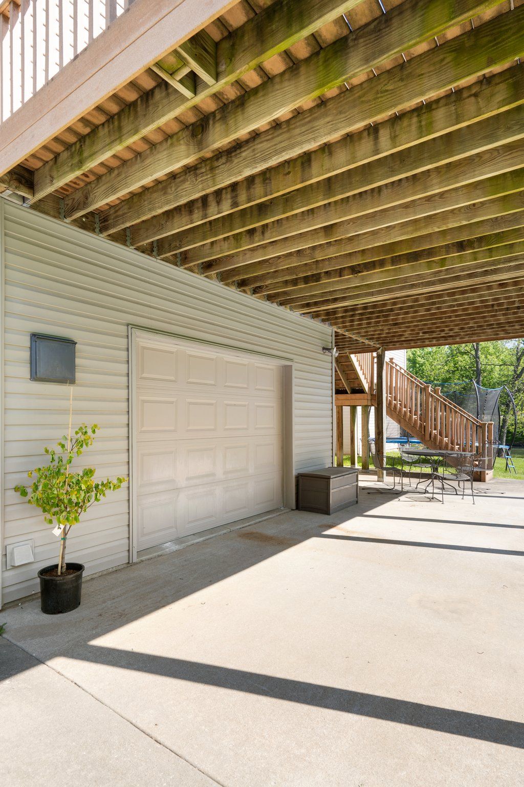 Exterior view: garage door under a wooden deck, concrete patio. A potted tree is visible.