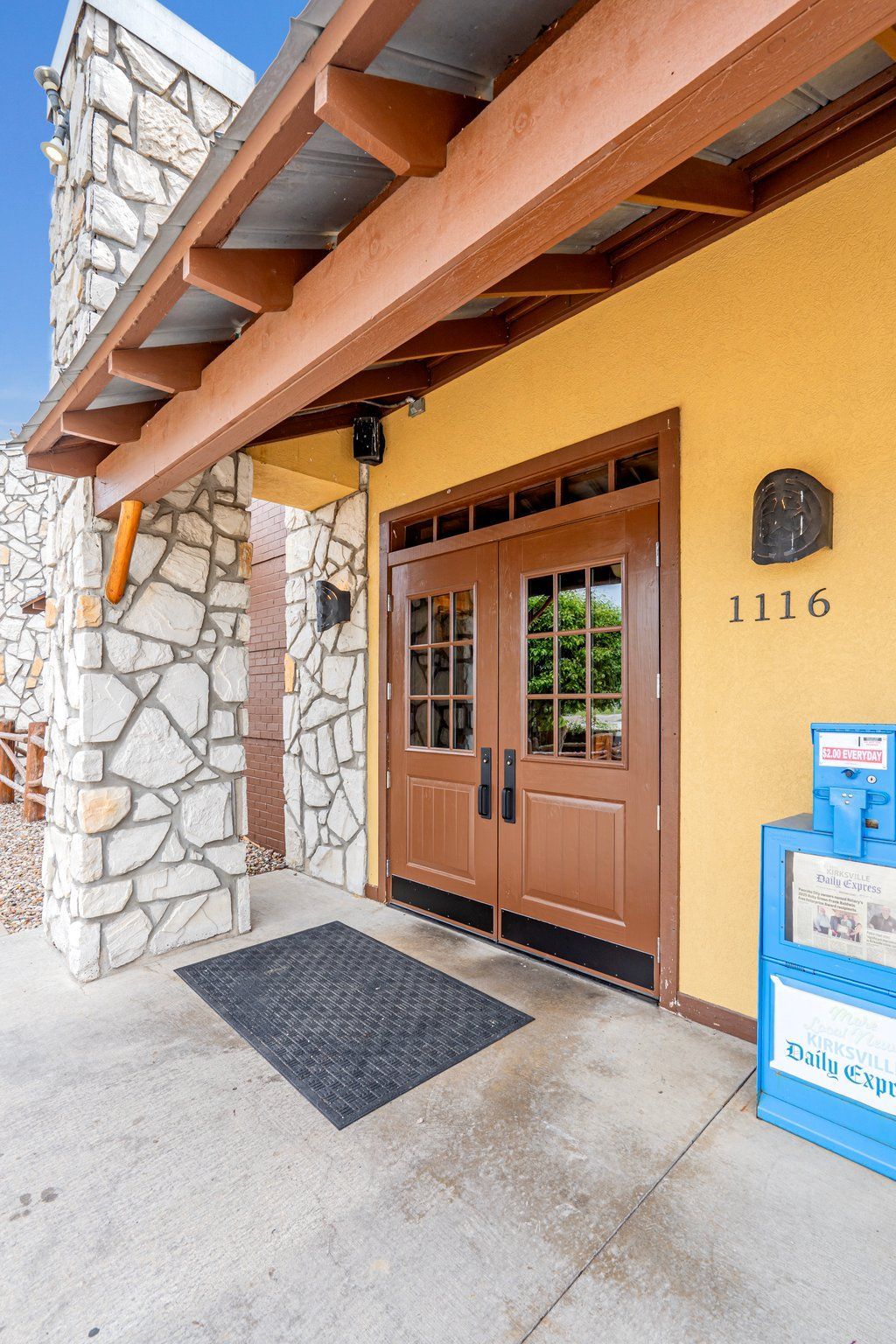Exterior view of a building with stone wall and brown double doors. Address on yellow stucco.