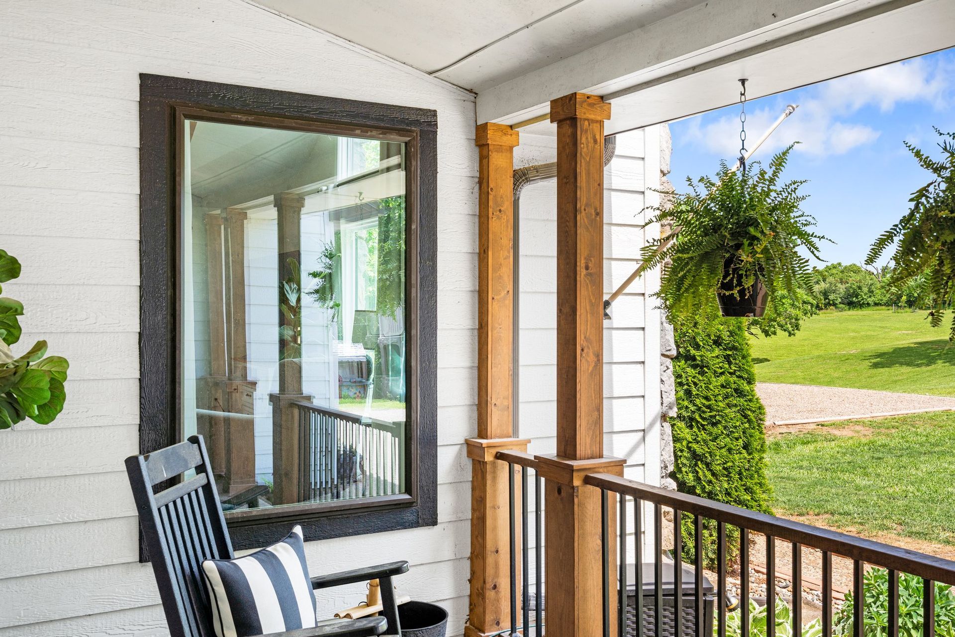Porch with rocking chair, mirror, wooden pillars, and hanging ferns, overlooking green field.