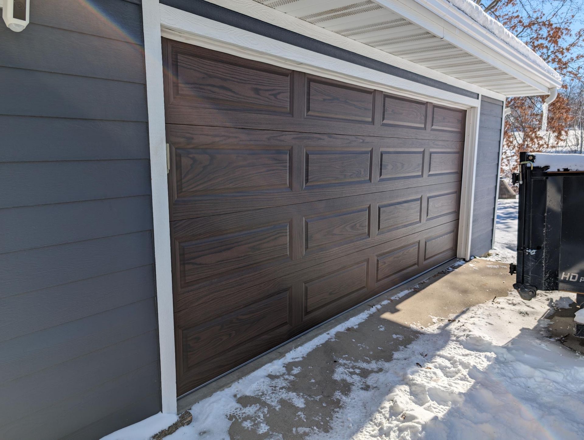 Dark brown wooden garage door on a blue-gray house with snow on the ground.