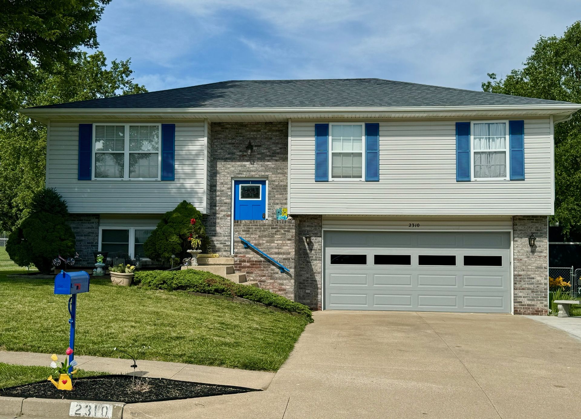 Two-story house with gray siding, blue accents, brick facade, and garage. Green lawn, blue mailbox, sunny day.