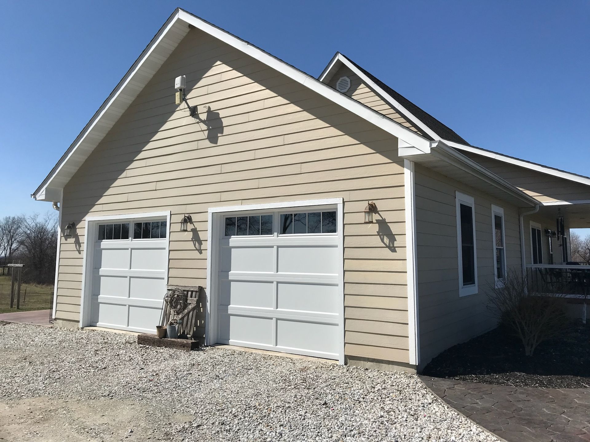 Tan-sided garage with two white garage doors, gravel driveway, and a glimpse of the connected house.