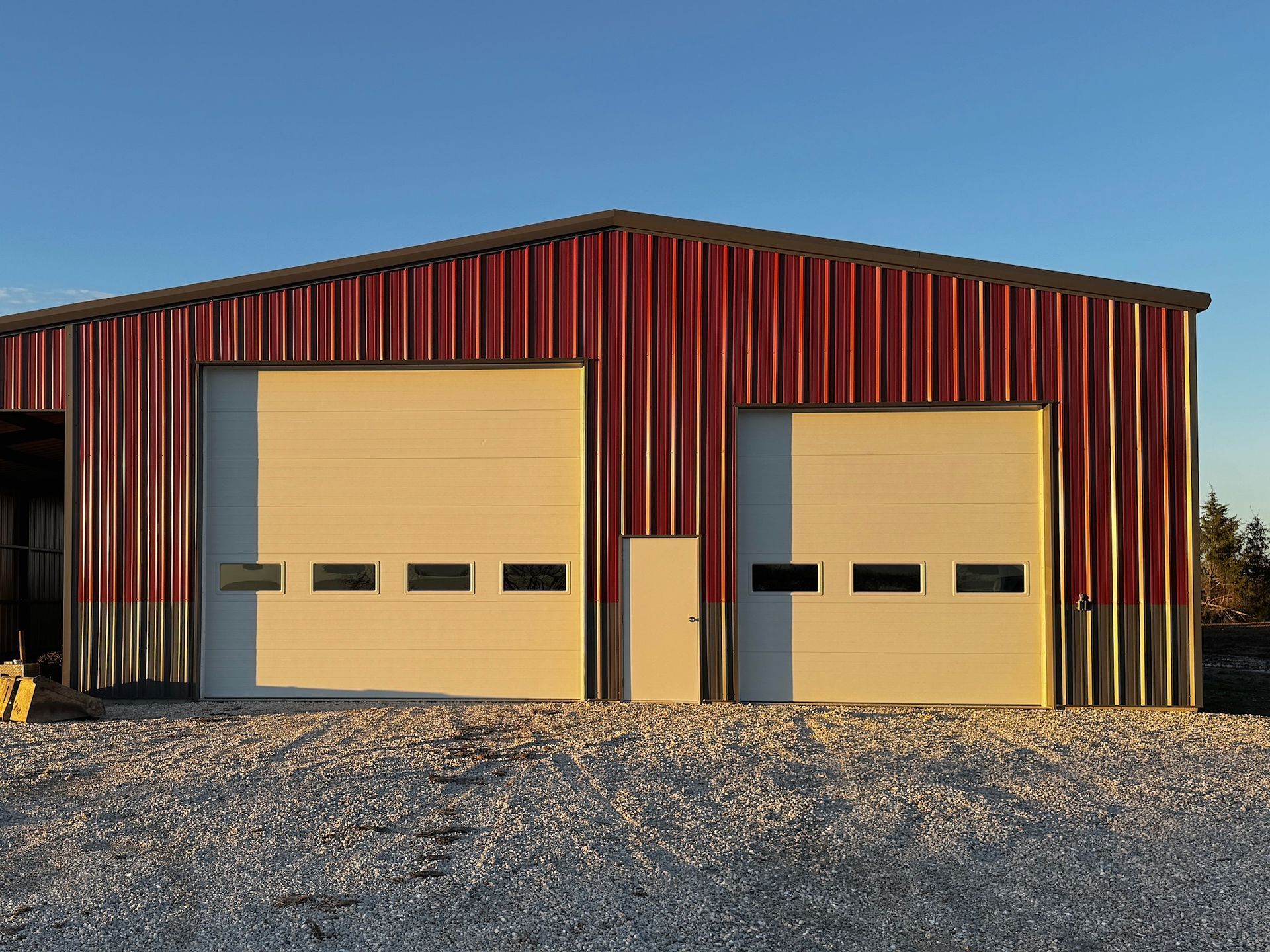 Red metal building with two garage doors and gravel driveway.