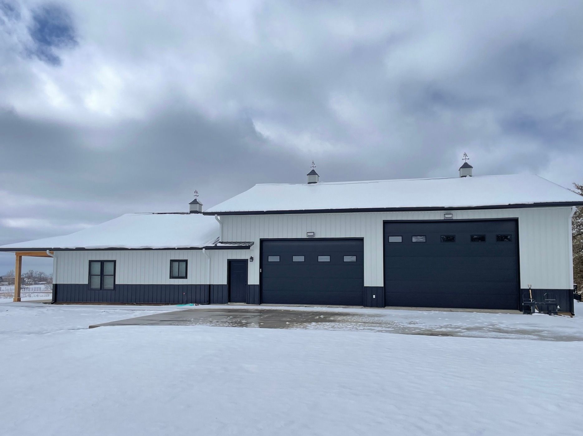 White and black barn-style building with snow-covered roof and two large garage doors. Gray sky.