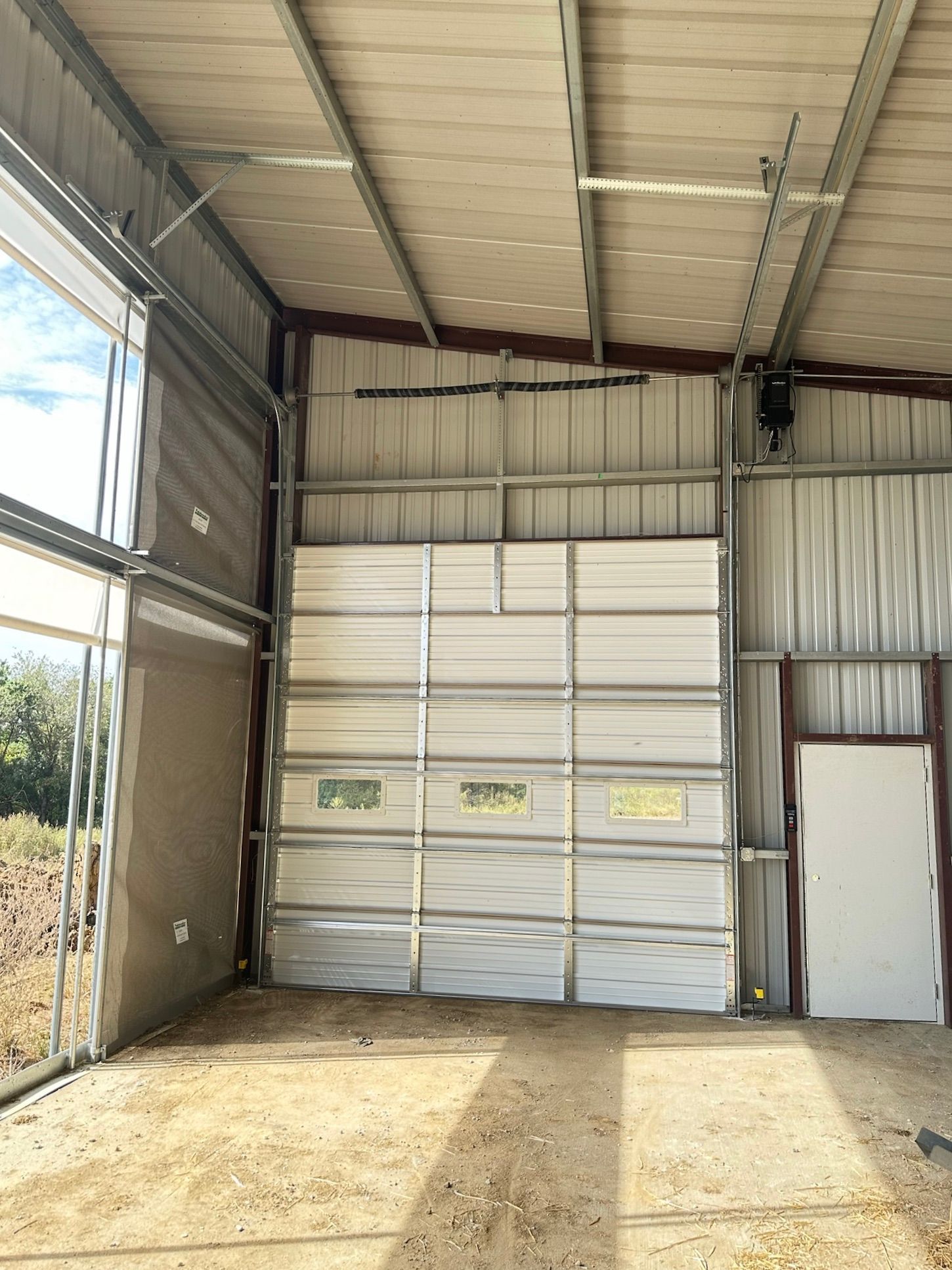 Interior of a metal building with a closed garage door, white side door, and open window to the left.