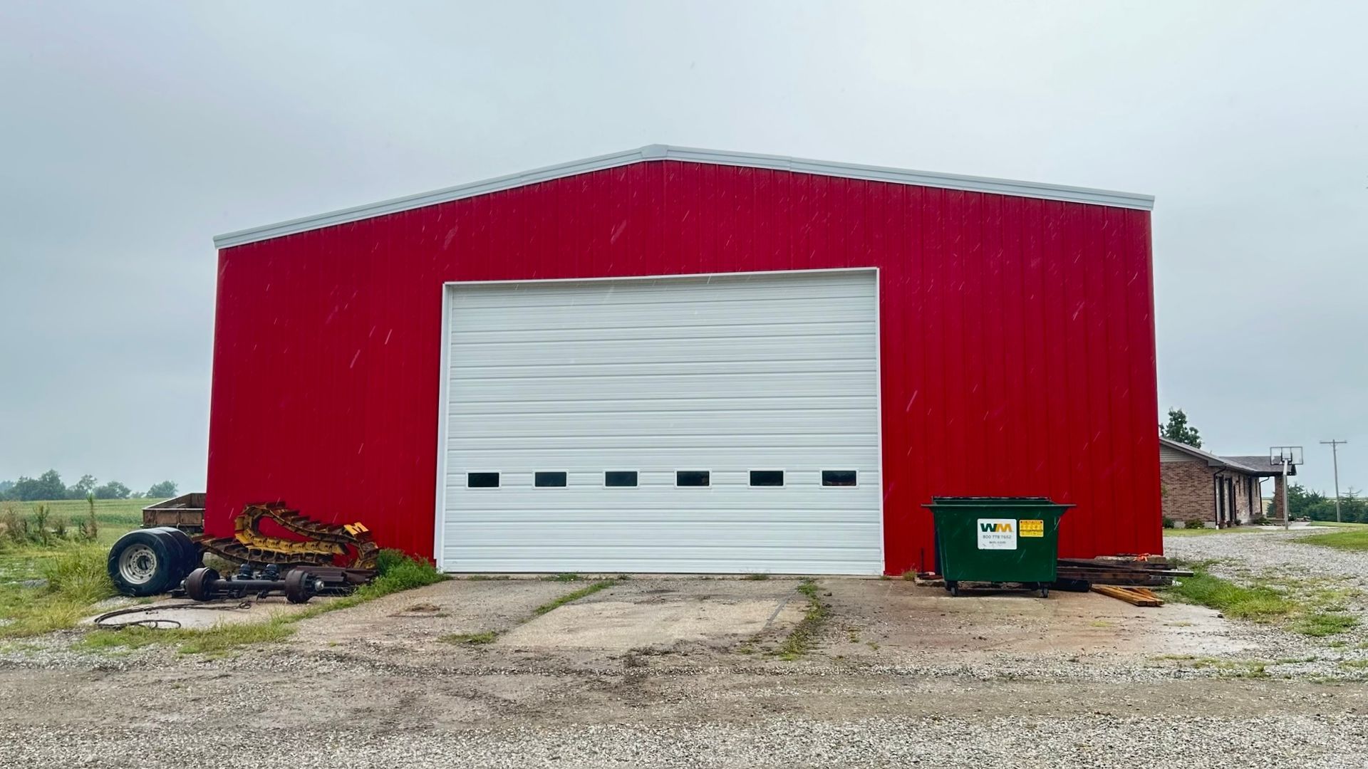 Red metal shed with a white garage door, a green dumpster, and farm equipment on a gravel lot.
