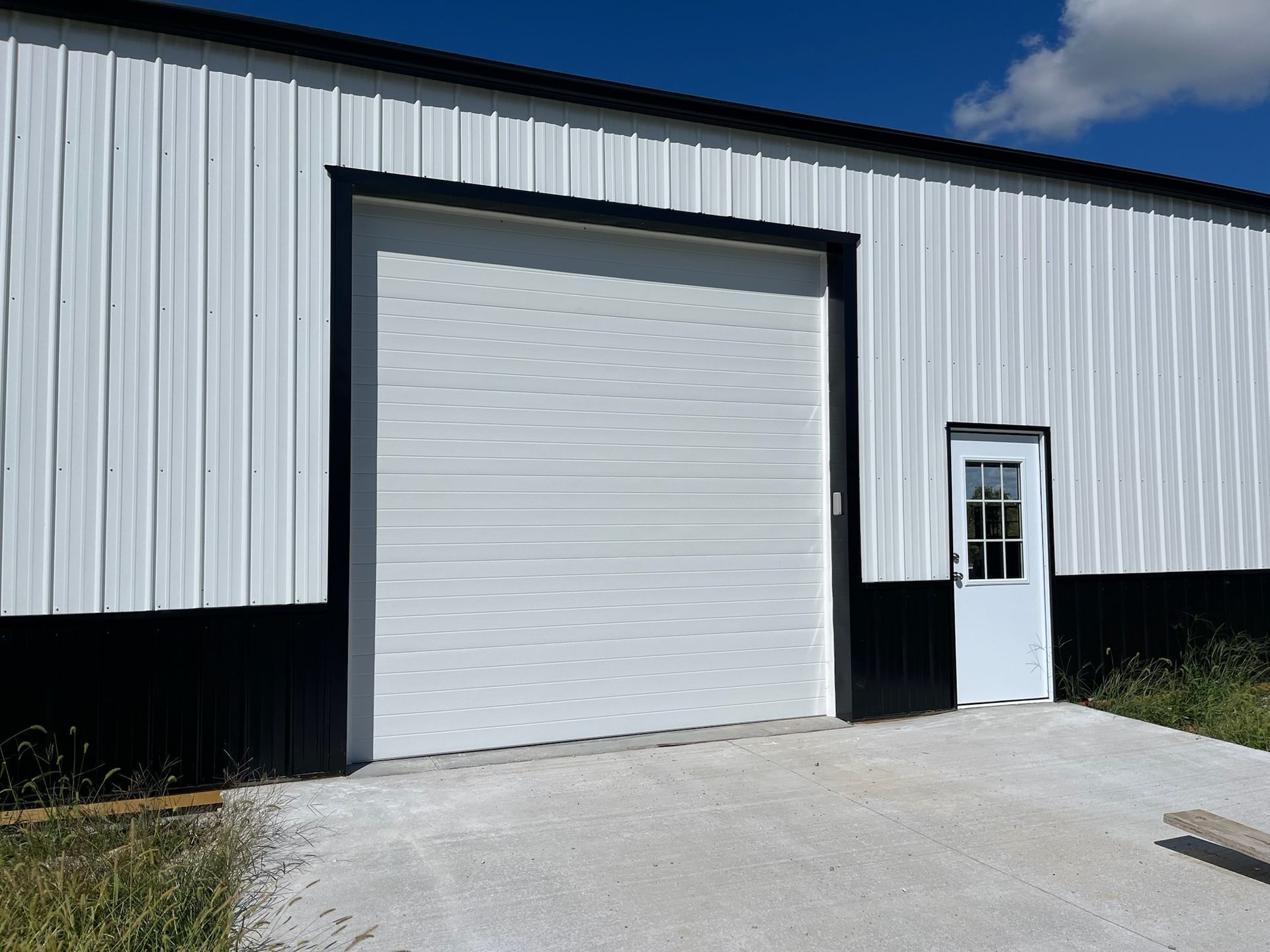White industrial building with a large garage door, small door, and concrete pad. Black trim.