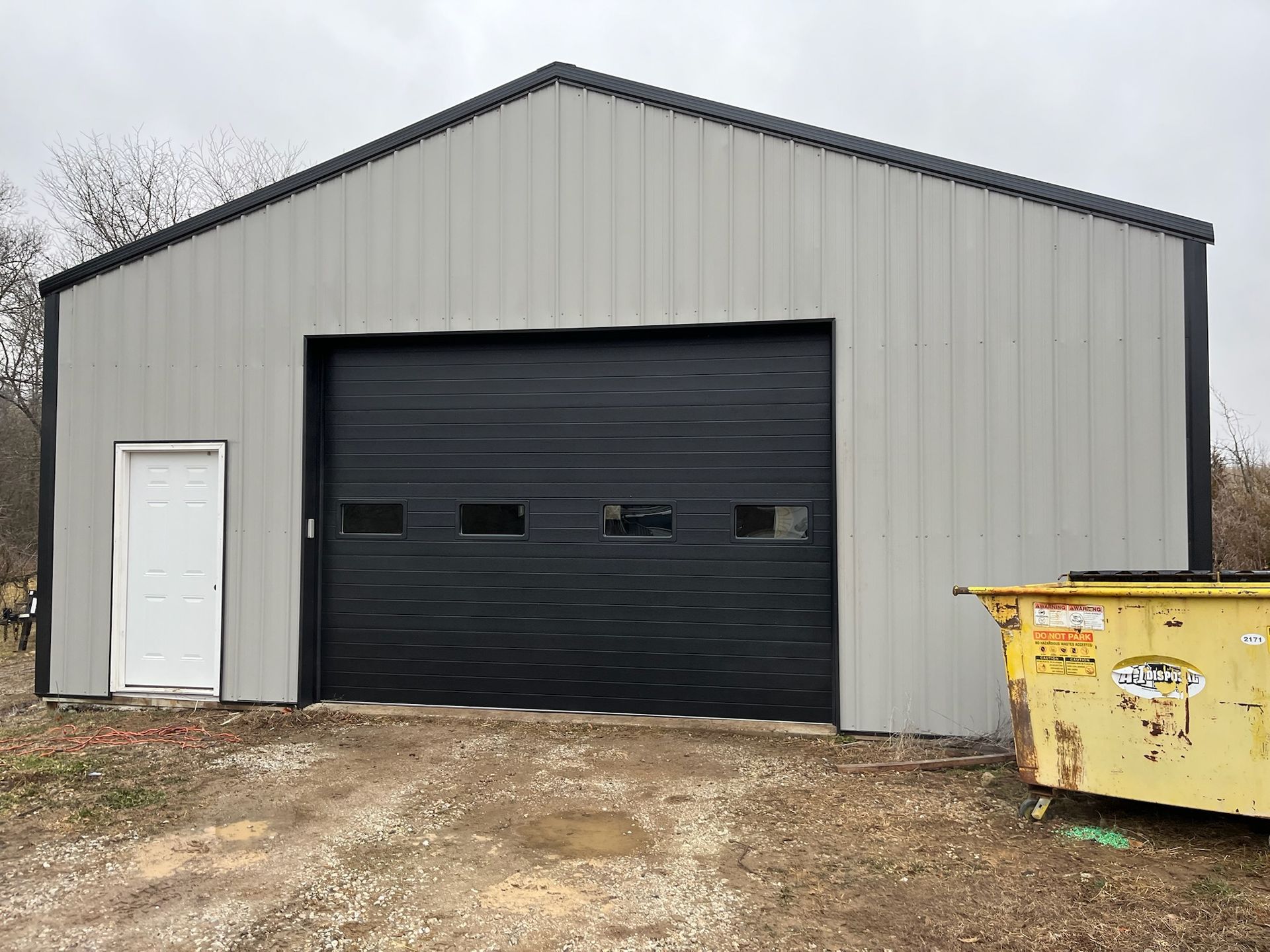 Gray metal building with a black garage door and a white door, with a yellow dumpster.