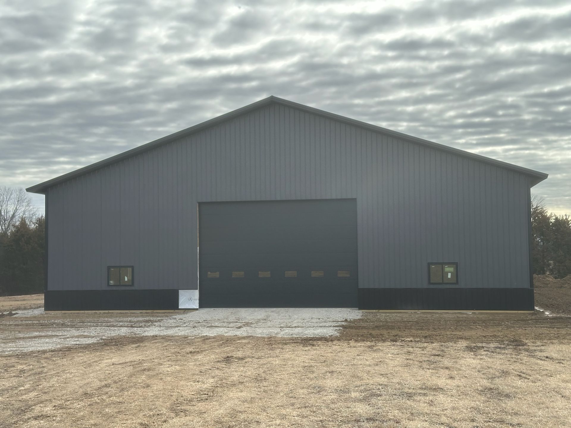 Dark gray metal building with large door, two small windows, gravel base, cloudy sky.