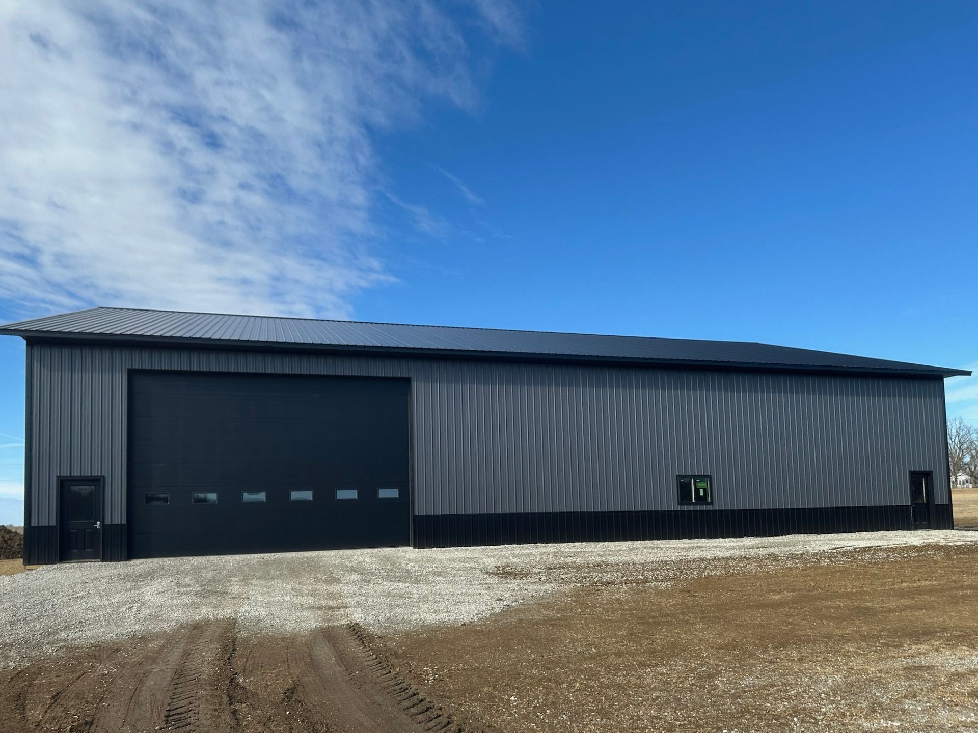 Gray metal building with large black overhead door, gravel drive, and blue sky.