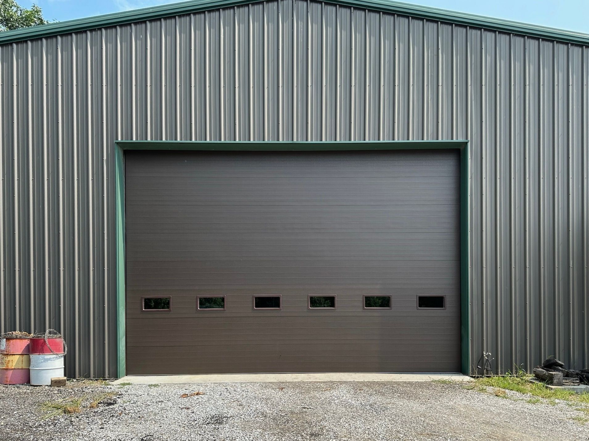 Brown garage door on a metal building with green trim. Gravel driveway, blue sky.
