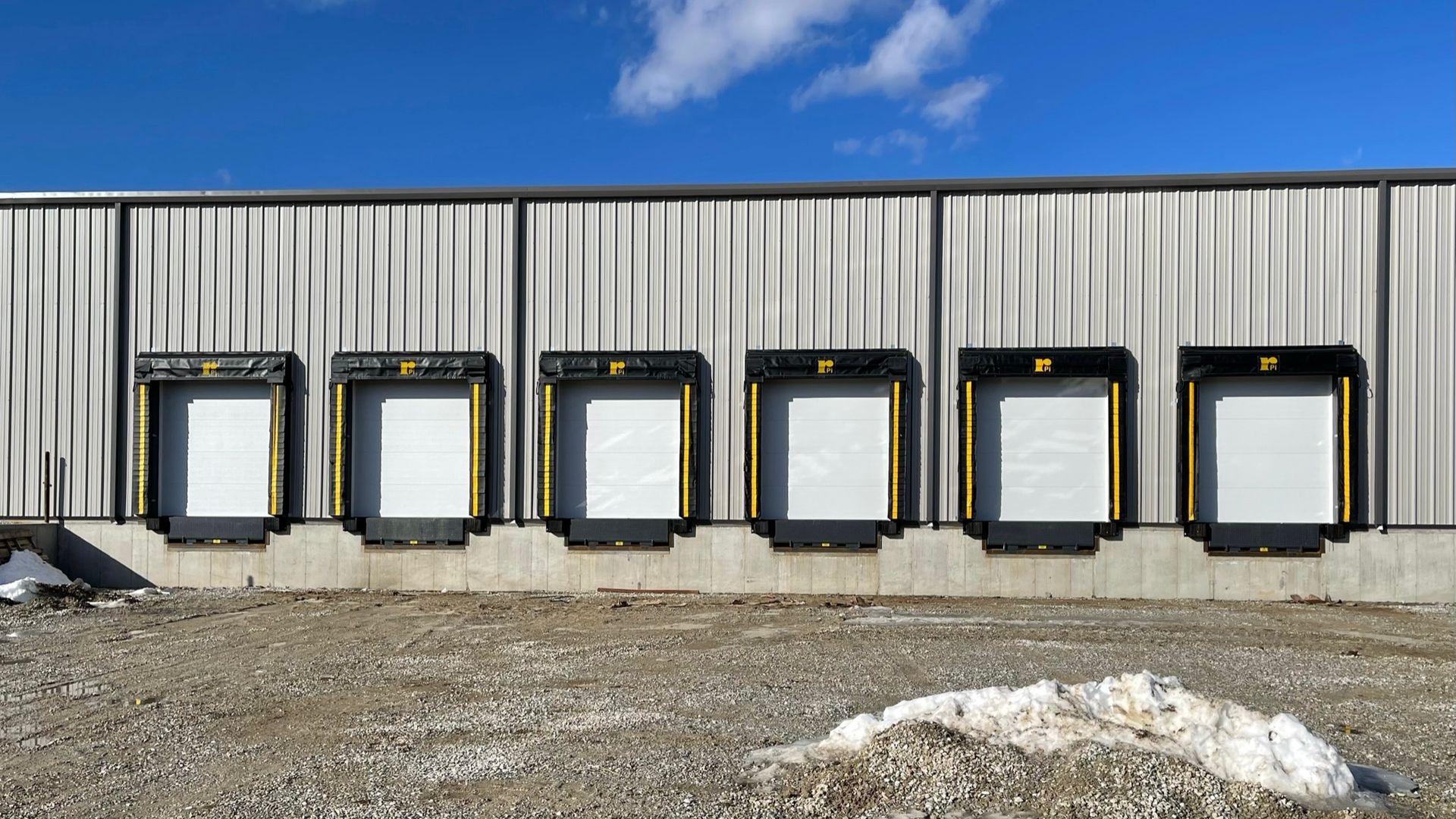 Exterior view of a building with six loading docks; white doors, black frames, yellow accents.
