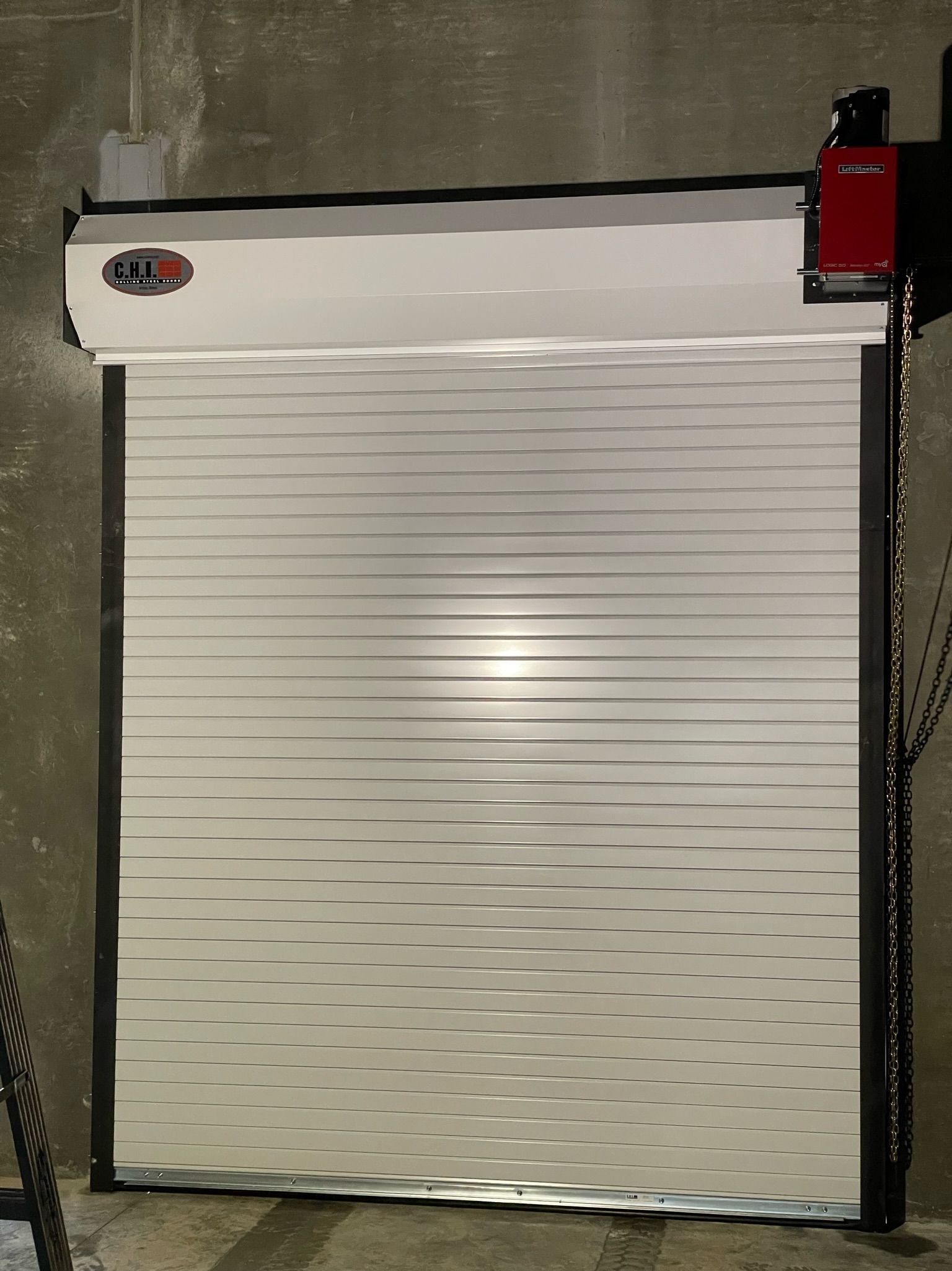 White, corrugated roll-up security door closed in a concrete setting with black vertical tracks and a red motor.