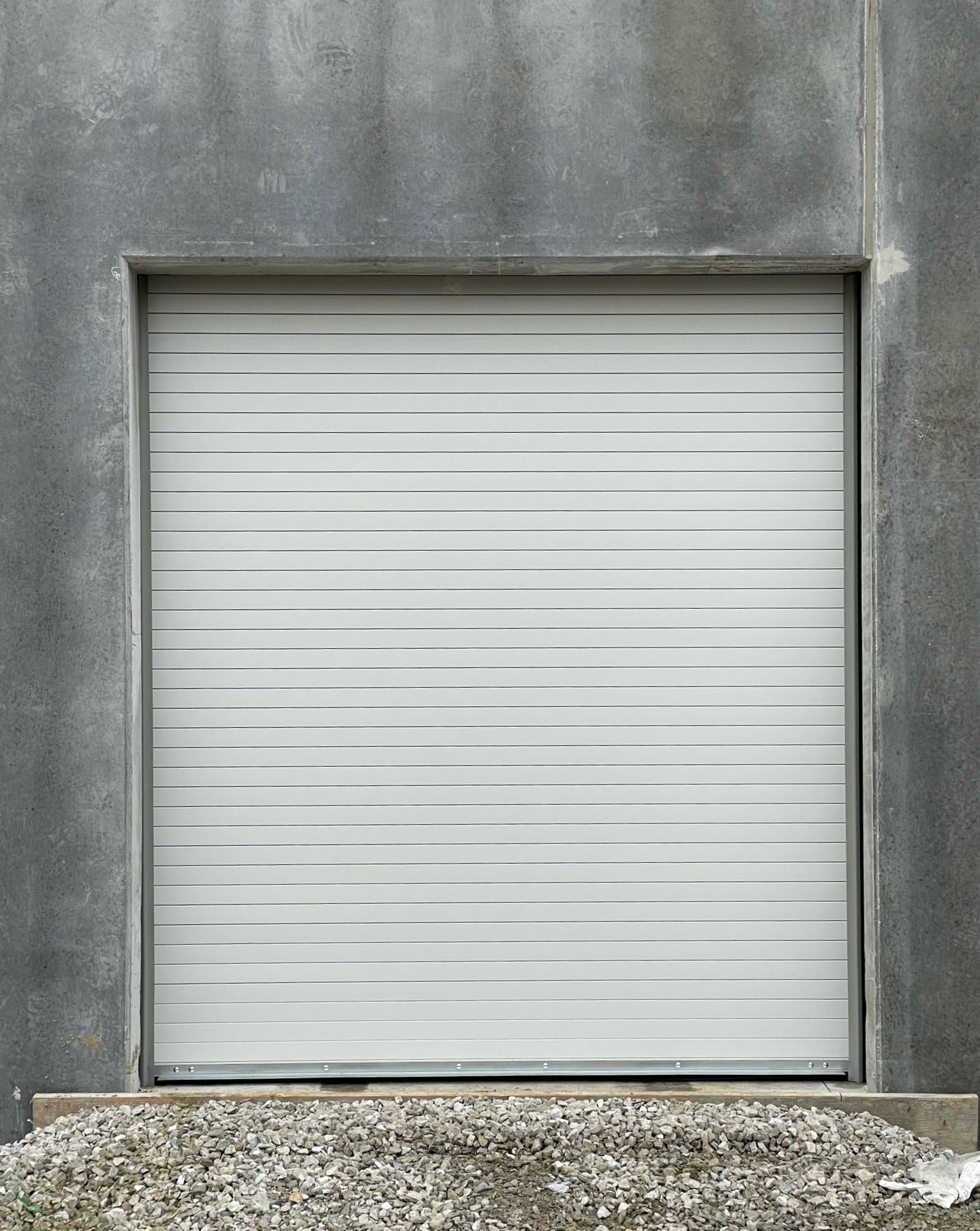 Gray roll-up door in a gray concrete building, set above gravel.