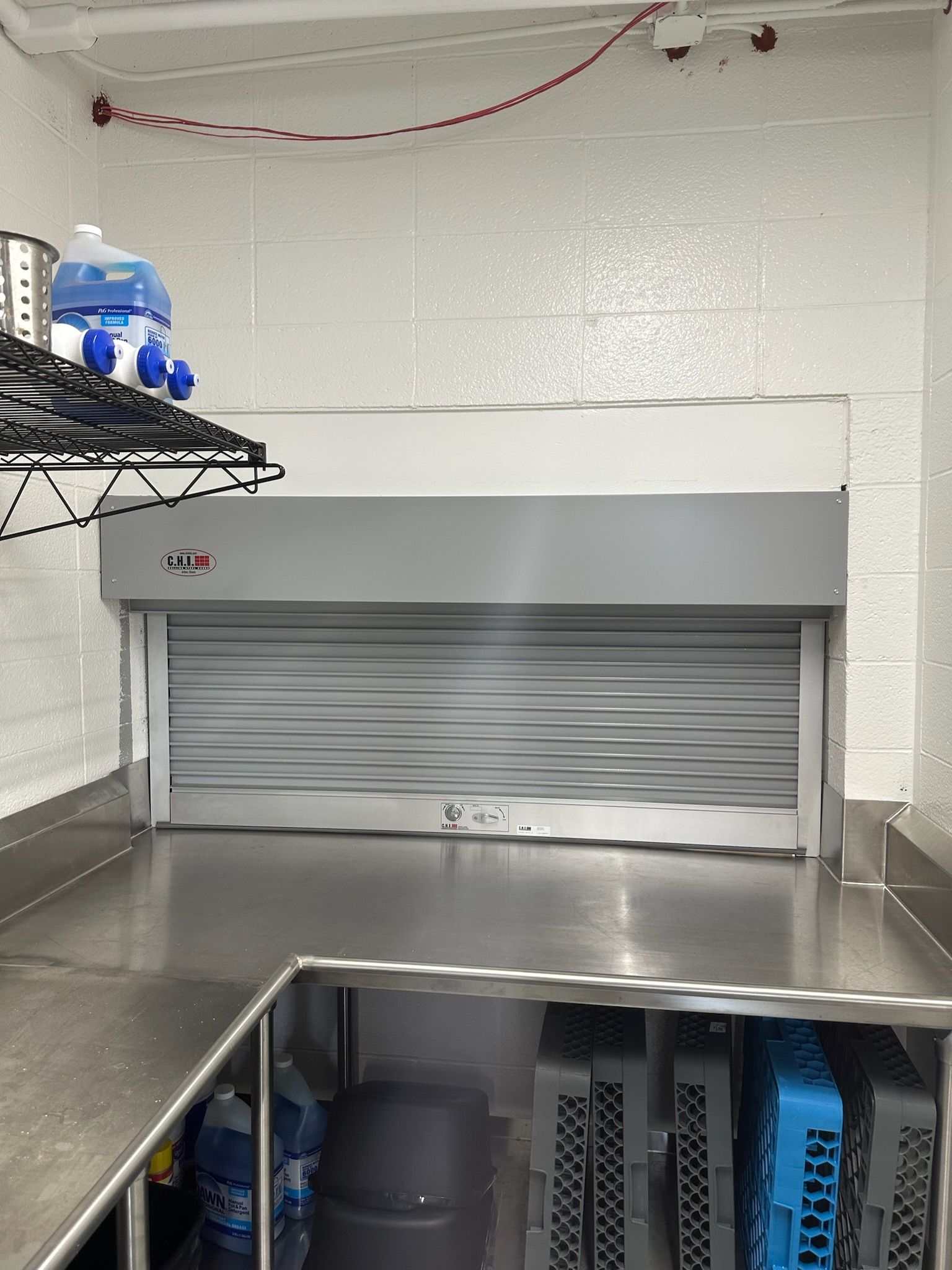 Stainless steel commercial kitchen with a closed rolling door, shelf with supplies, and dish racks.