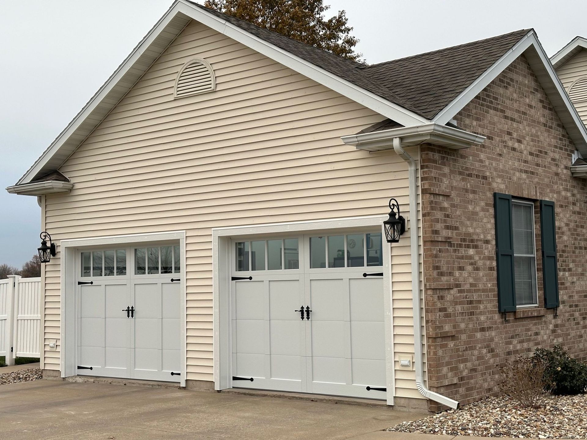 Two-car garage with white doors, tan siding, and brick facade. Black accents and outdoor lights.