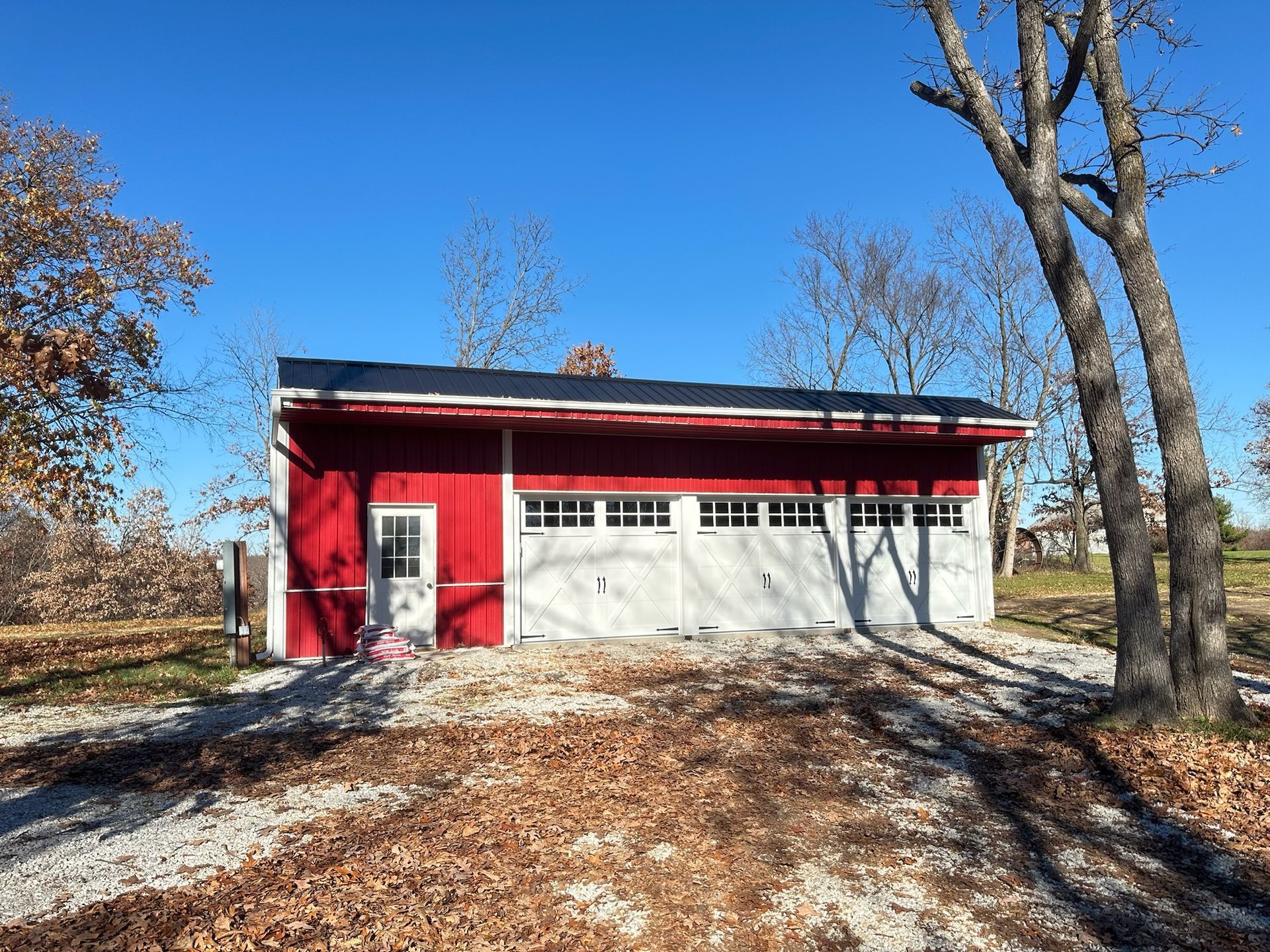 Red and white garage with black roof, three garage doors, and a small door on a gravel driveway under a blue sky.