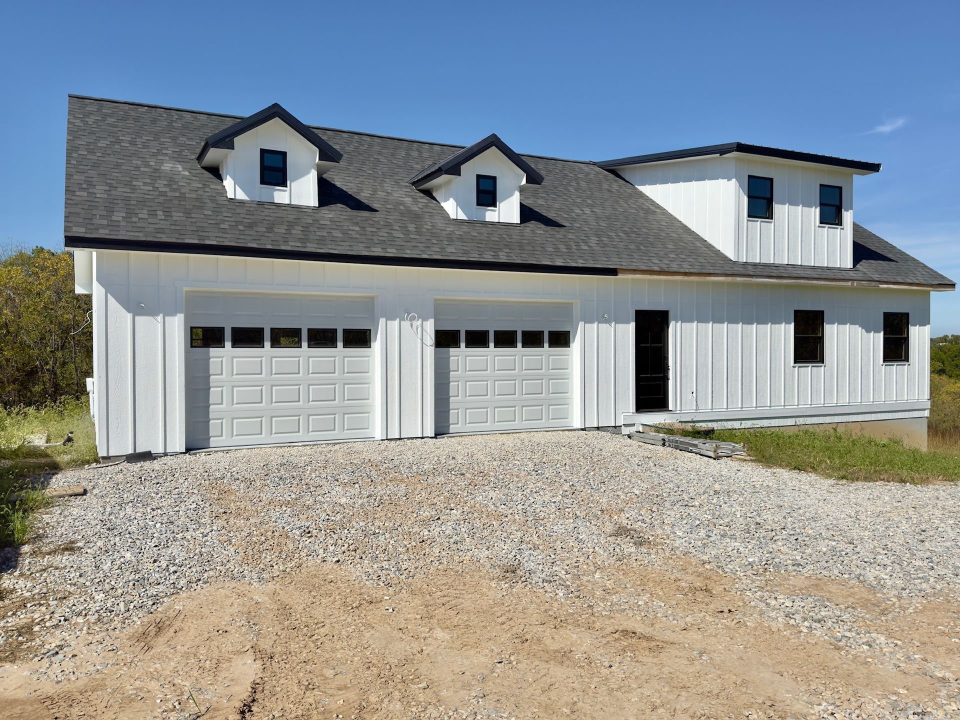 White two-car garage with black trim, three dormers, and gravel driveway under a blue sky.