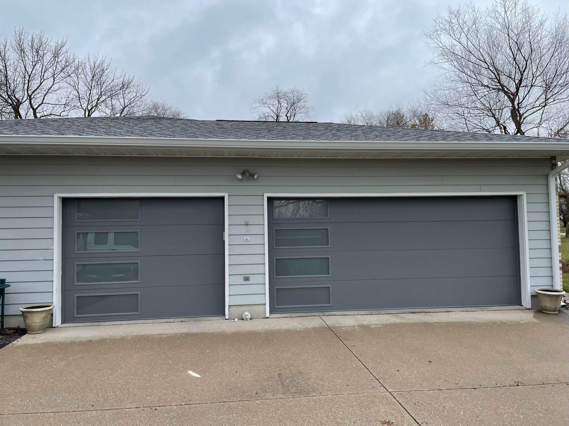Two-car garage with gray doors, windows, and light gray siding. Cloudy sky, two trees.