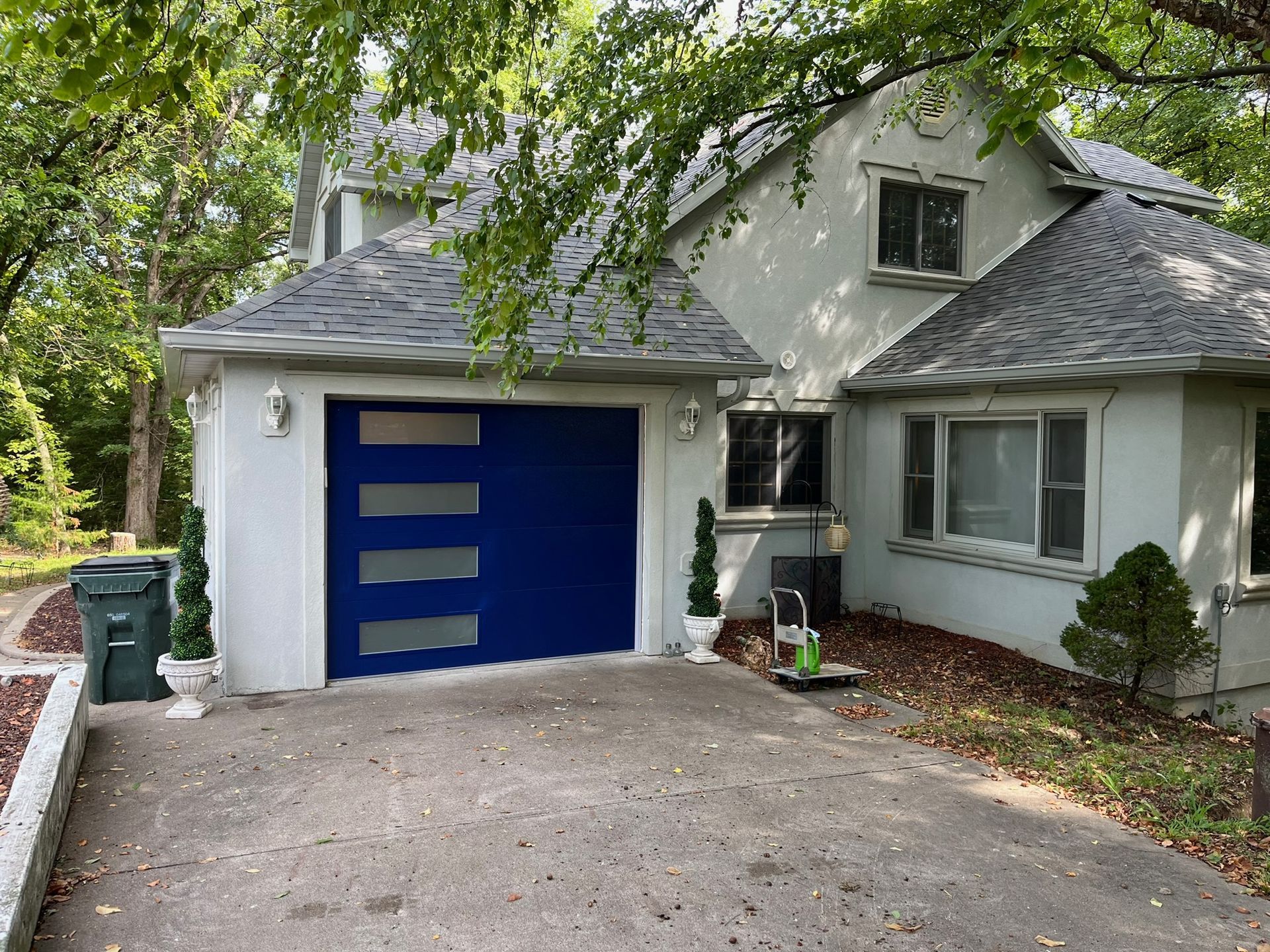 Two-story house with blue garage door, white stucco exterior, and gray roof. Driveway leads up to it.