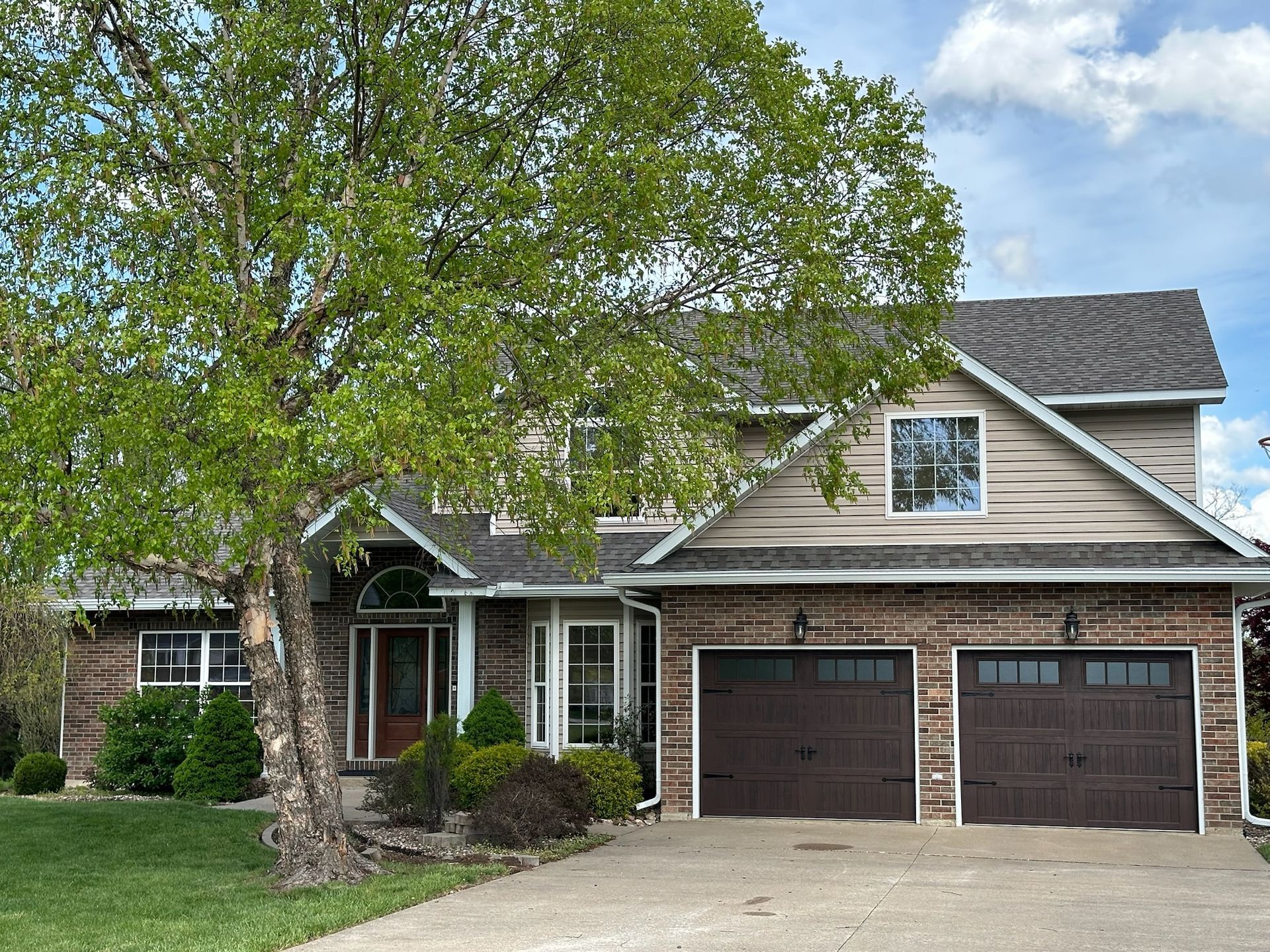 Two-story house with brown garage doors, brick and tan siding, and a tree in front.
