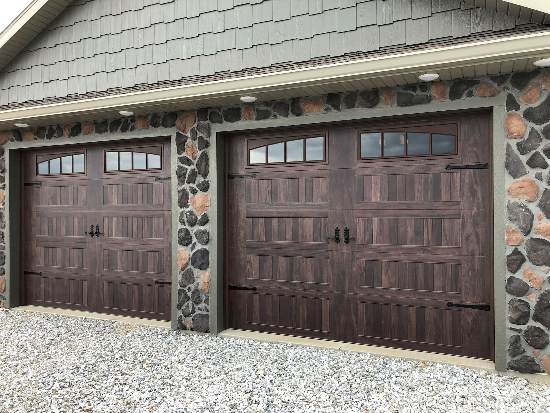 Two dark brown garage doors with decorative windows, flanked by stone walls, under a gray roof.