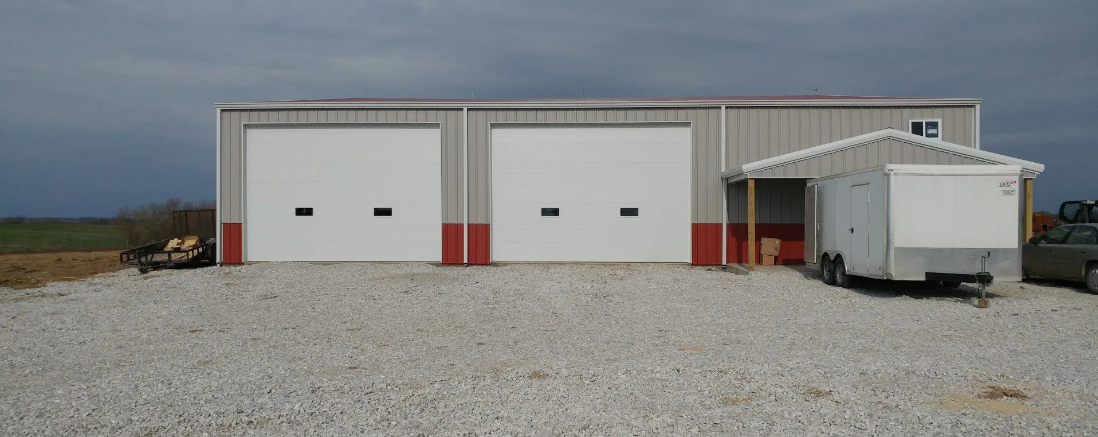 A gray metal building with two garage doors and a white trailer on a gravel lot under a cloudy sky.